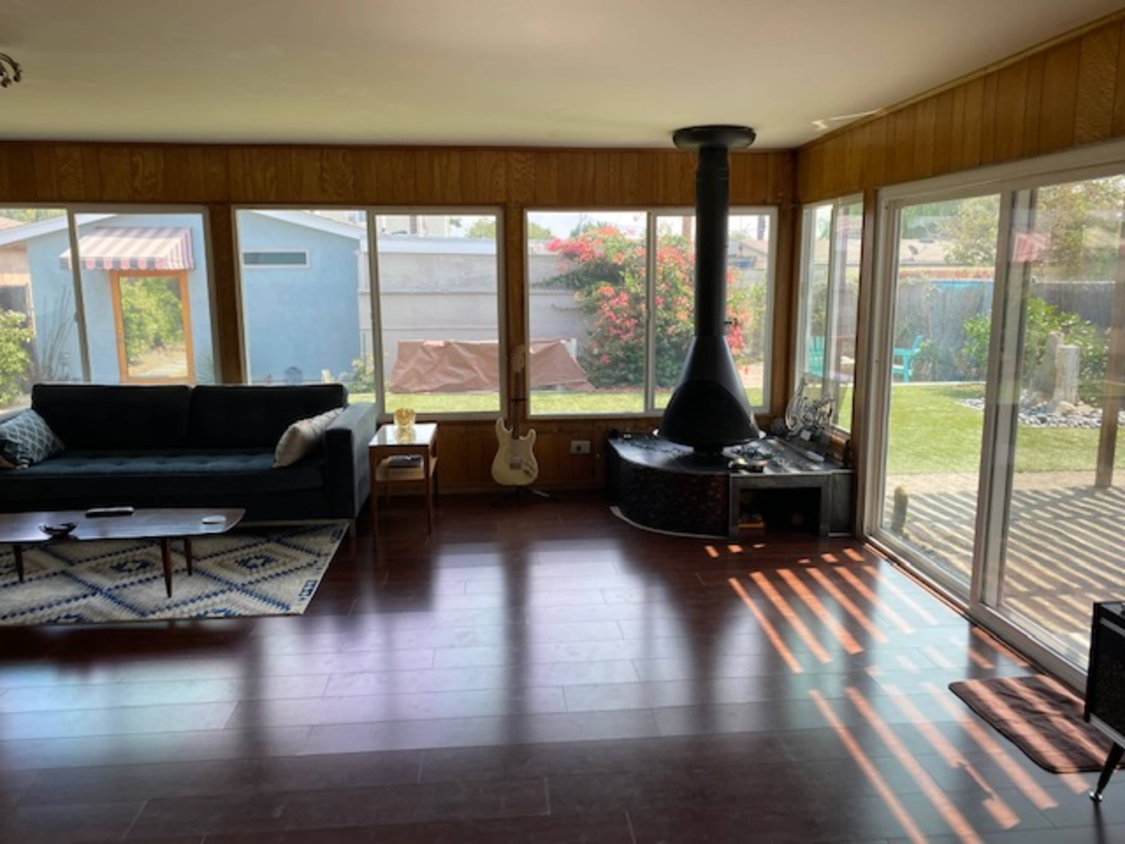 A sunny living room with a dark sofa, a coffee table, a guitar, and a modern chimney surrounded by large windows that overlook a green backyard.