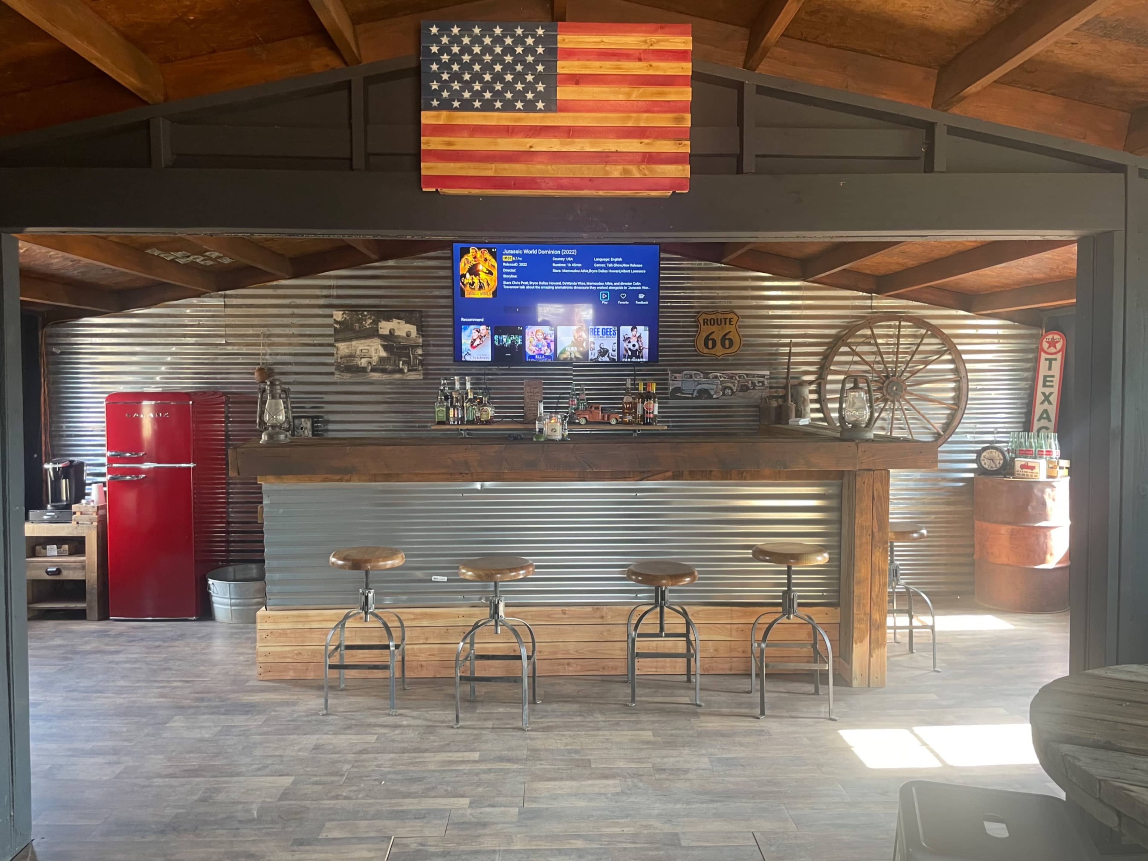 A rustic bar area with a corrugated metal wall, a large television mounted above the bar, a vintage red refrigerator, and several wooden bar stools.