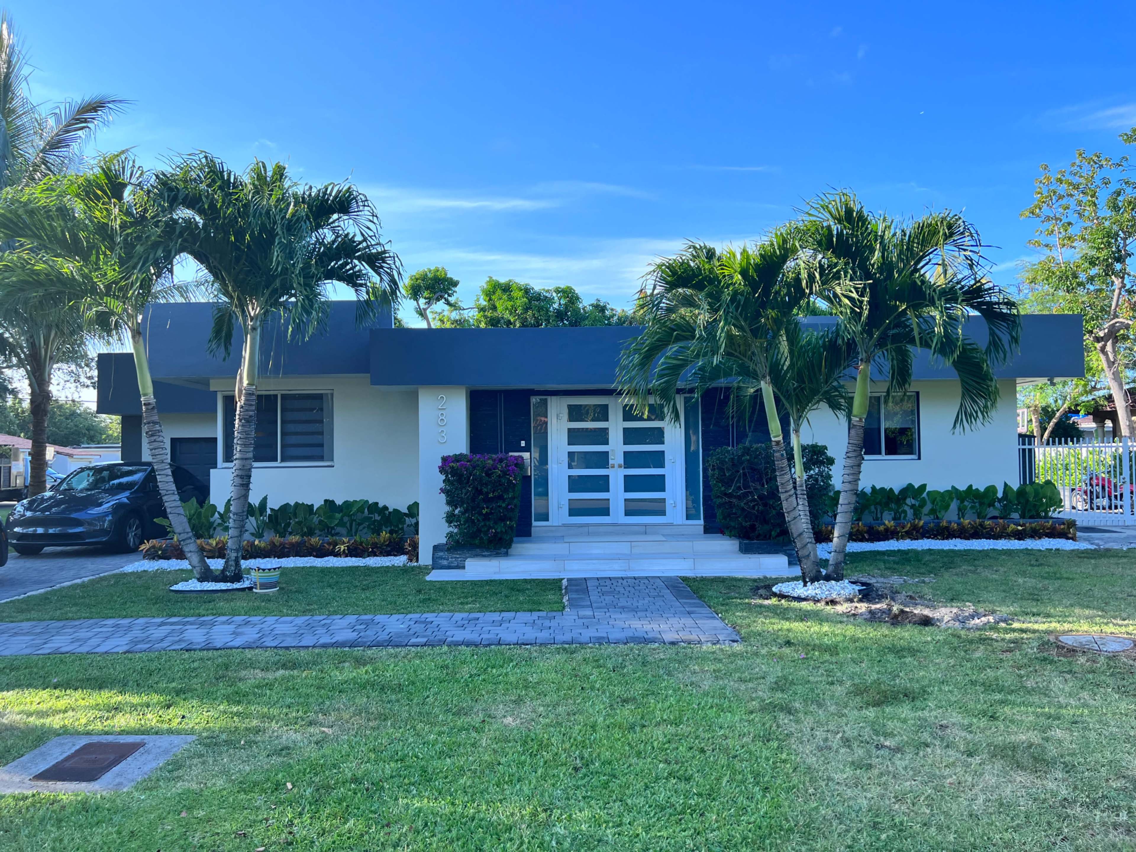 The image shows a modern single-story house with a landscaped front yard, bordered by palm trees and a stone pathway.