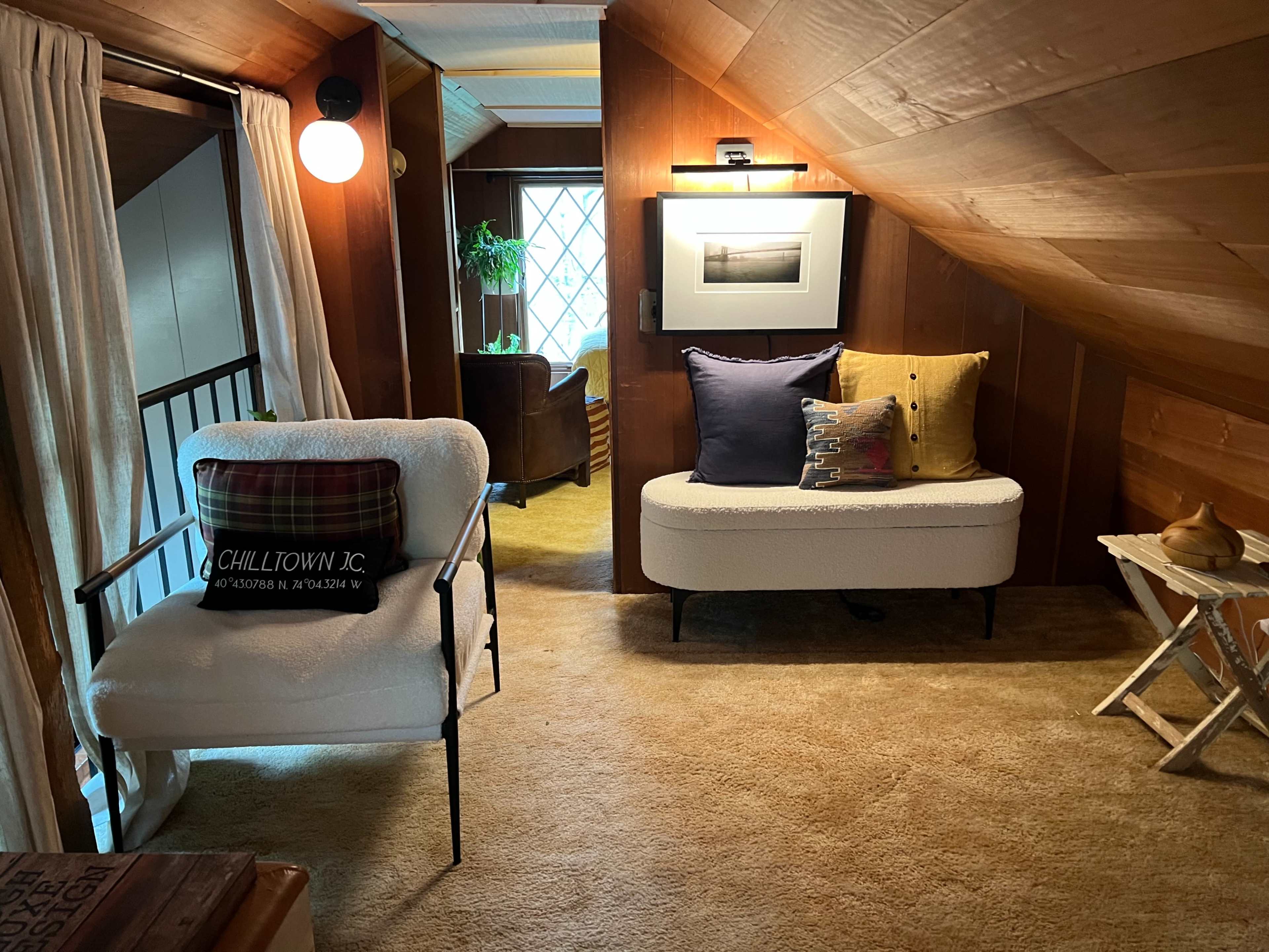 The image shows a cozy, warmly lit attic space featuring a small seating area with a chair and a sofa, as well as a decorative side table and framed artwork on the walls.