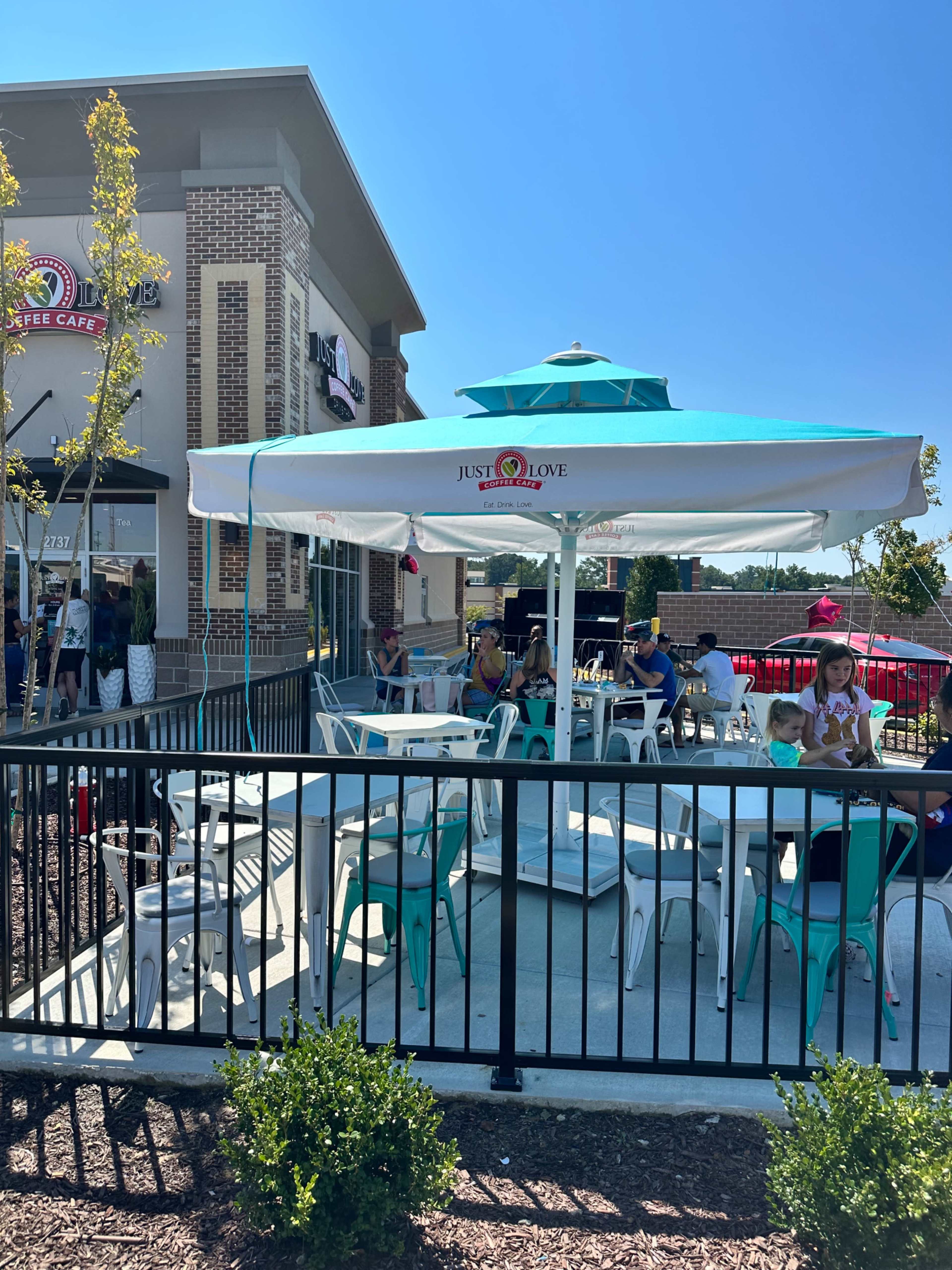 A patio area with white tables and turquoise chairs is situated outside a café, shaded by a large umbrella.