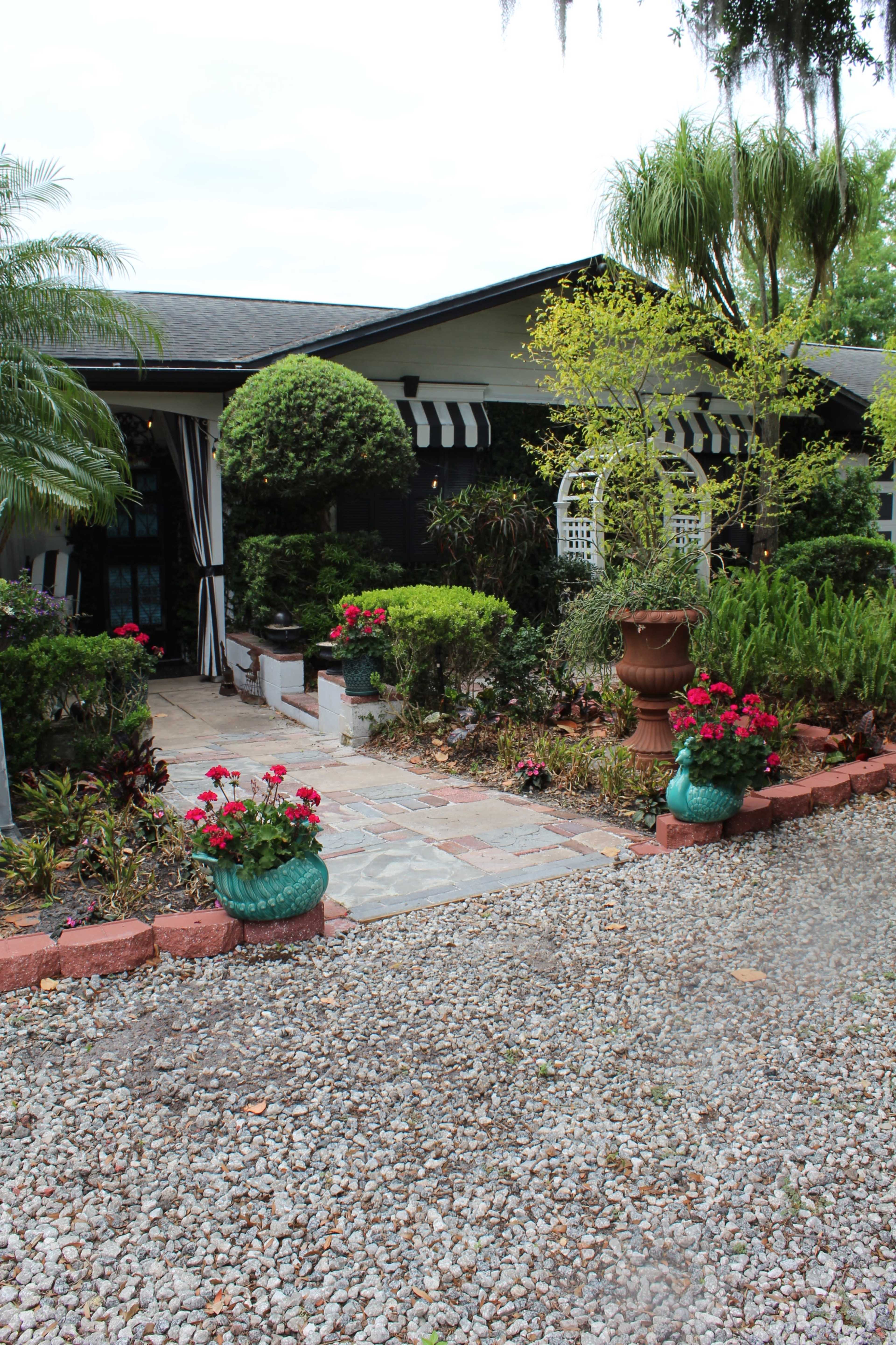 The image shows a landscaped front yard with a stone pathway leading to a house, surrounded by flower beds and decorative potted plants.