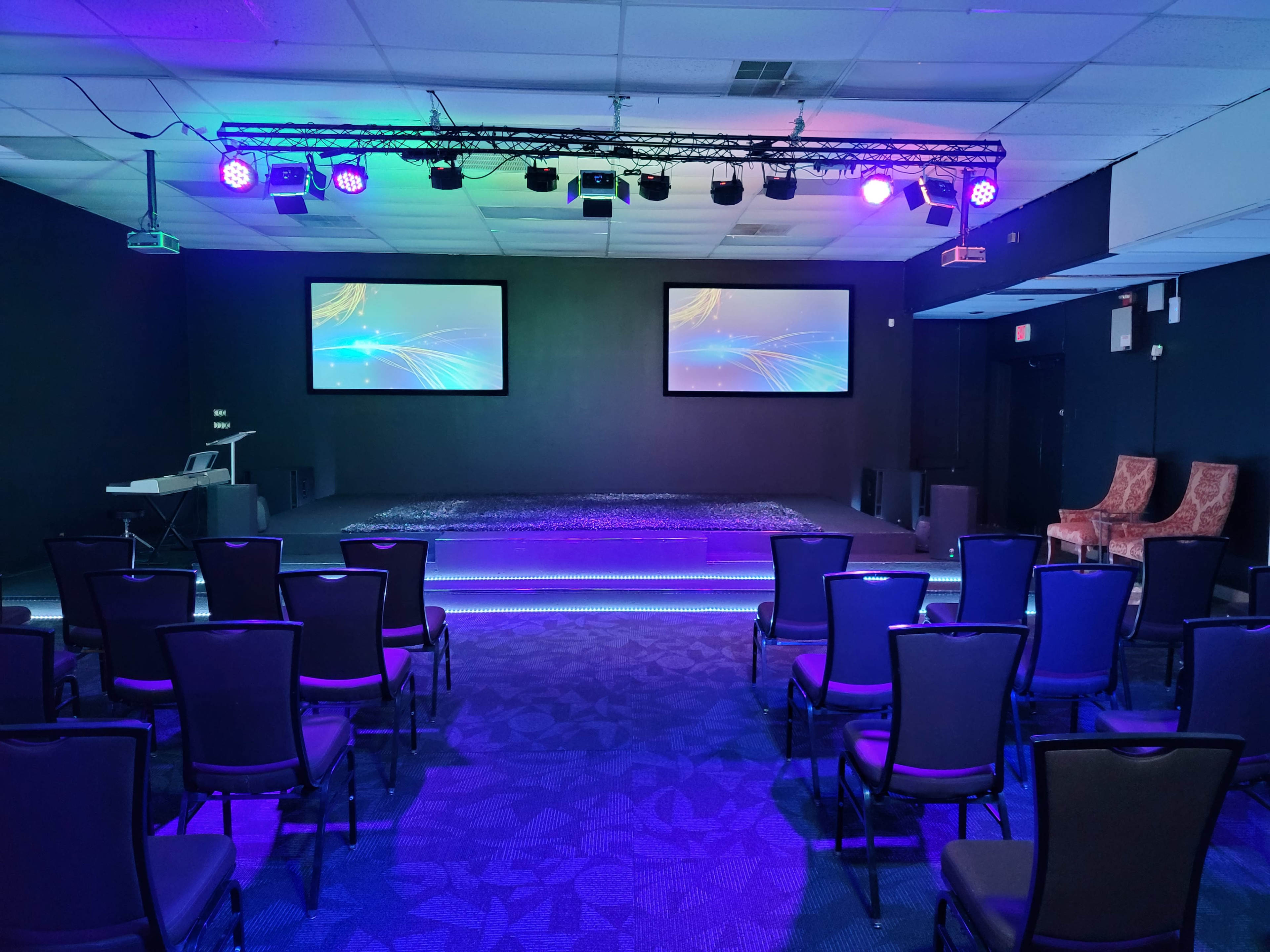 A dimly lit room set up for a presentation, featuring rows of black chairs facing a stage with two screens at the front and colorful lighting.