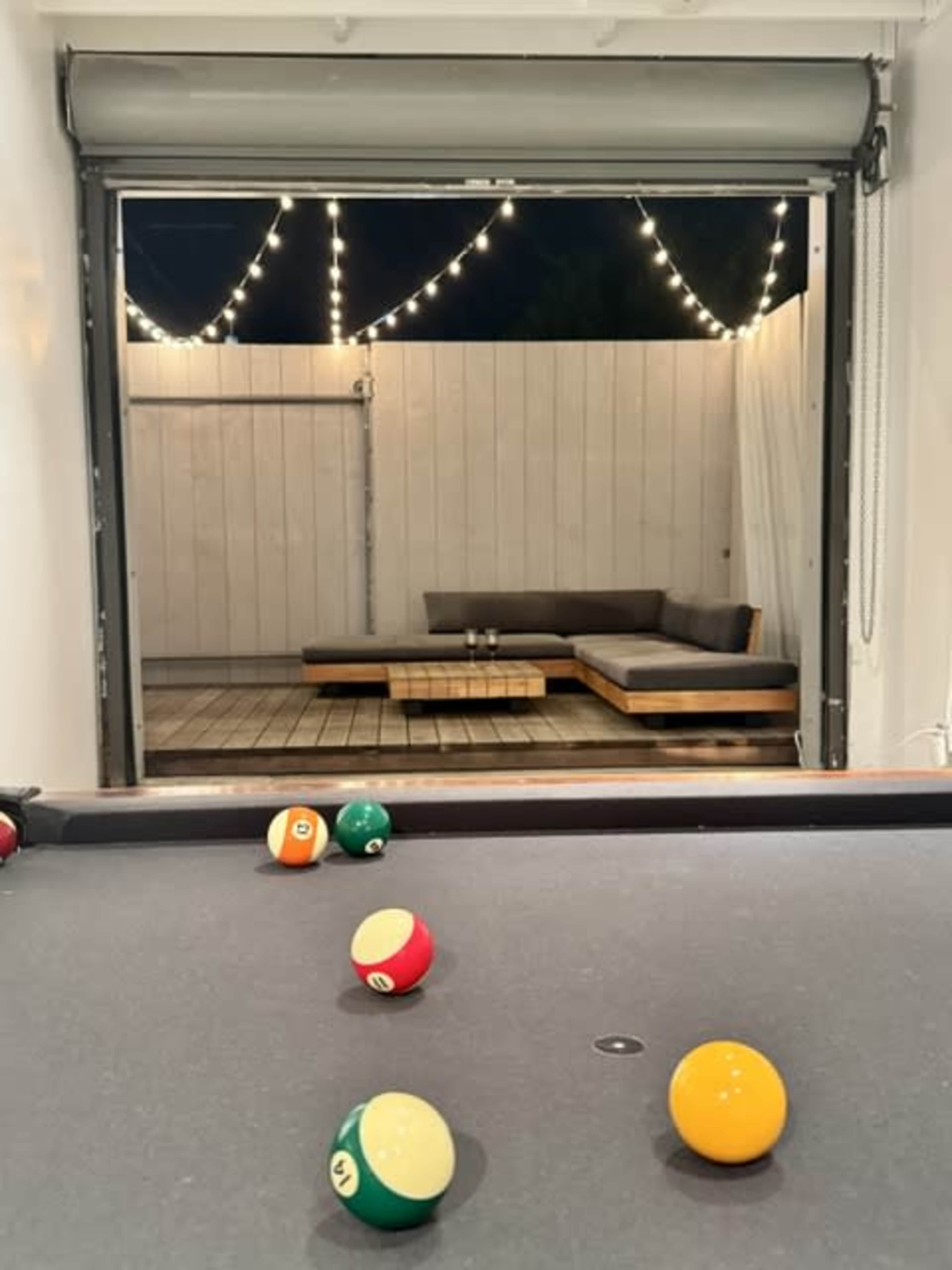 A pool table is in the foreground, with colorful balls scattered across it, while a cozy seating area illuminated by string lights is visible in the background through an open wall.