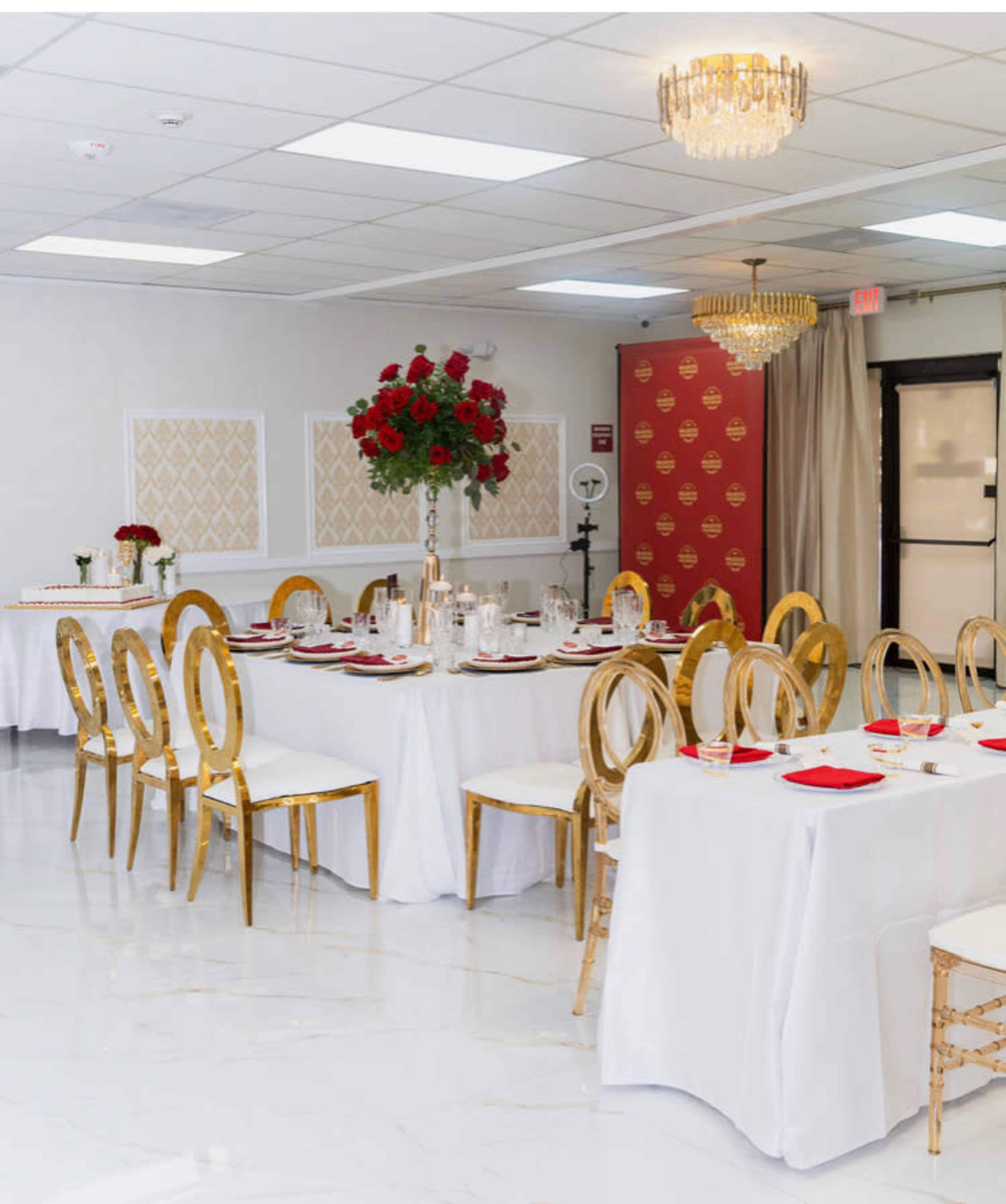 A banquet hall features elegantly set dining tables with white tablecloths, gold chairs, and floral centerpieces.