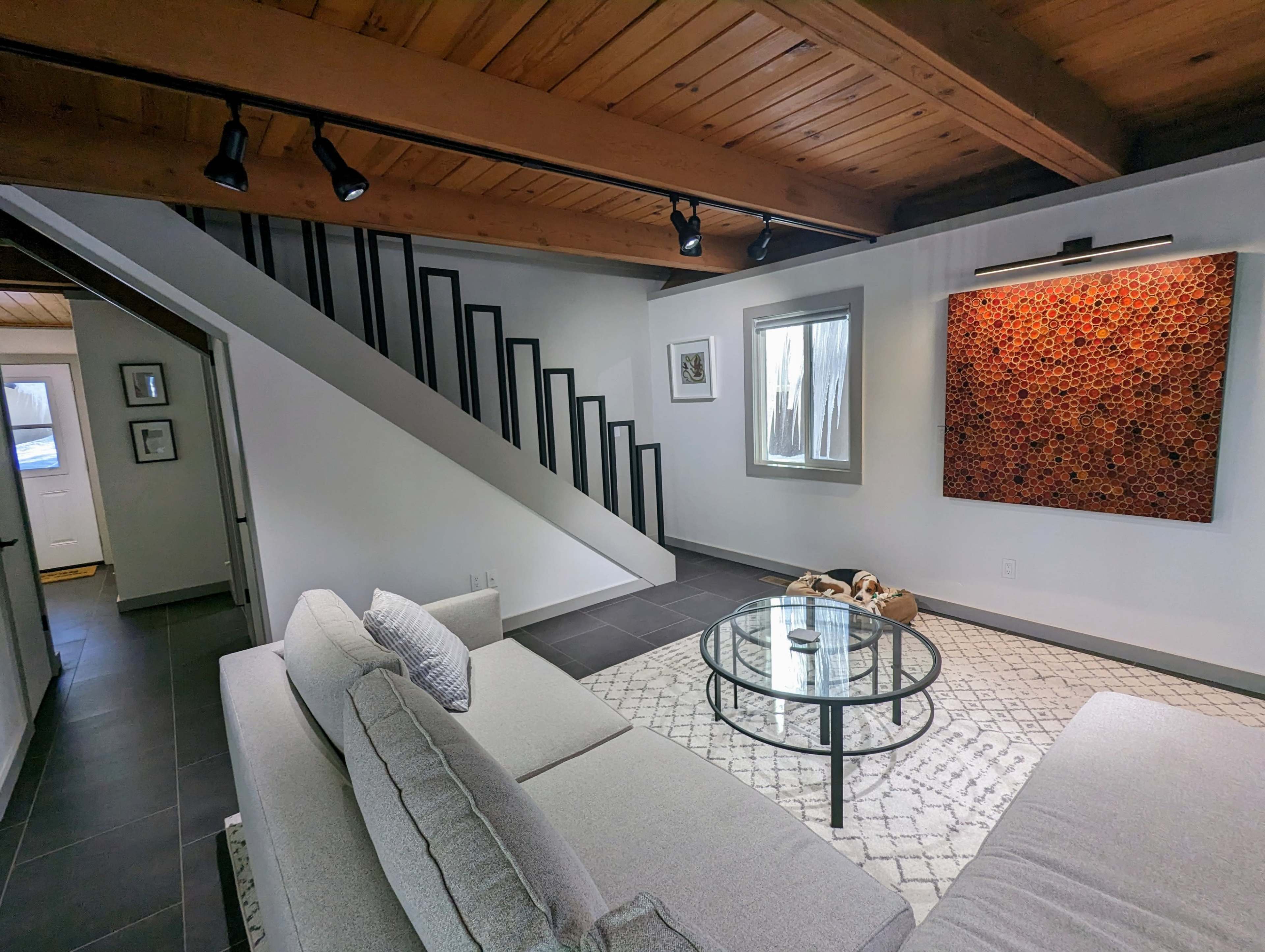 A modern living room features a sectional sofa, a round glass coffee table, and a large artwork on the wall, with a staircase visible in the background.