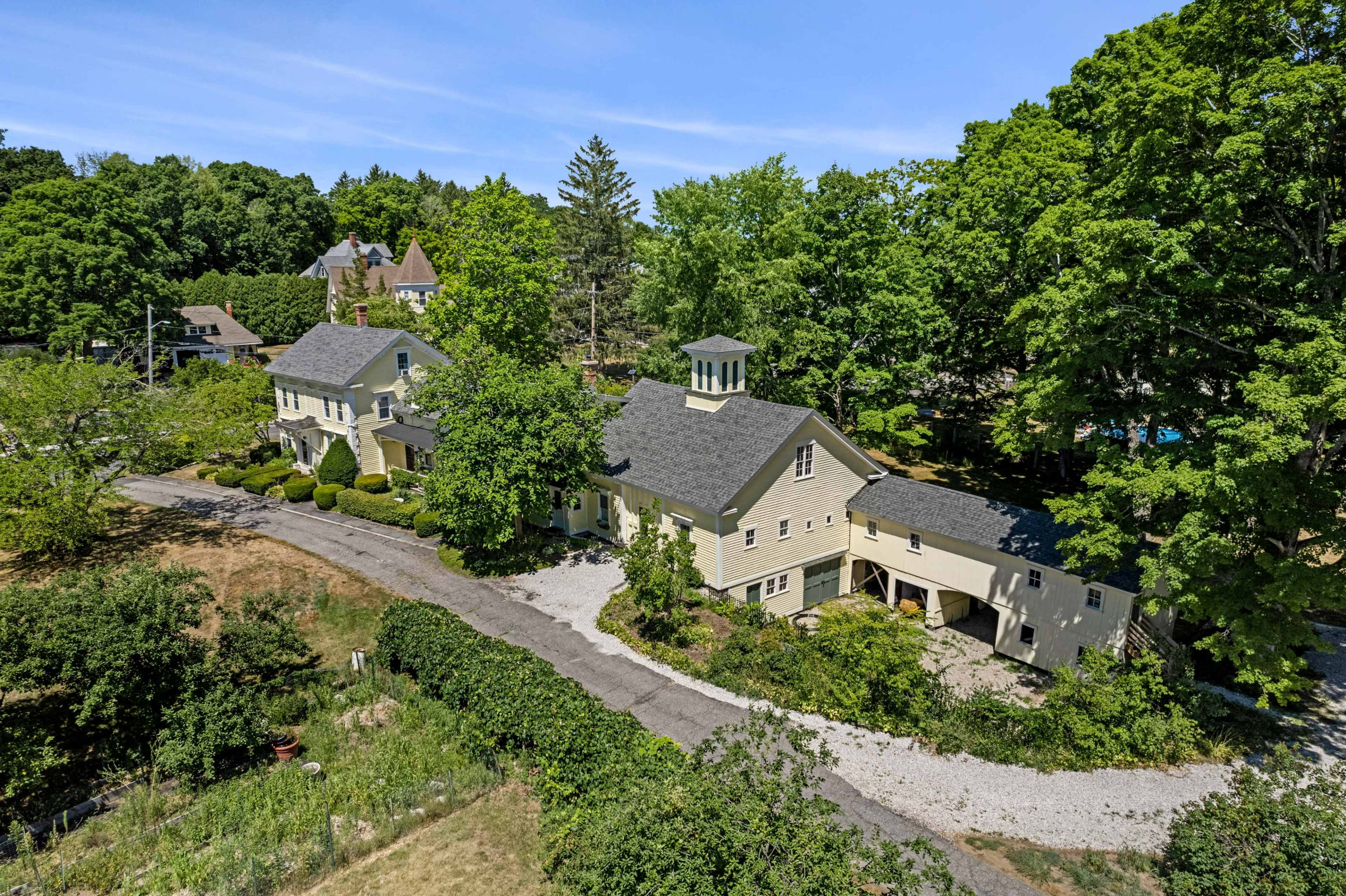A large, two-story house with a gray roof and a cupola, surrounded by green trees and a gravel driveway.