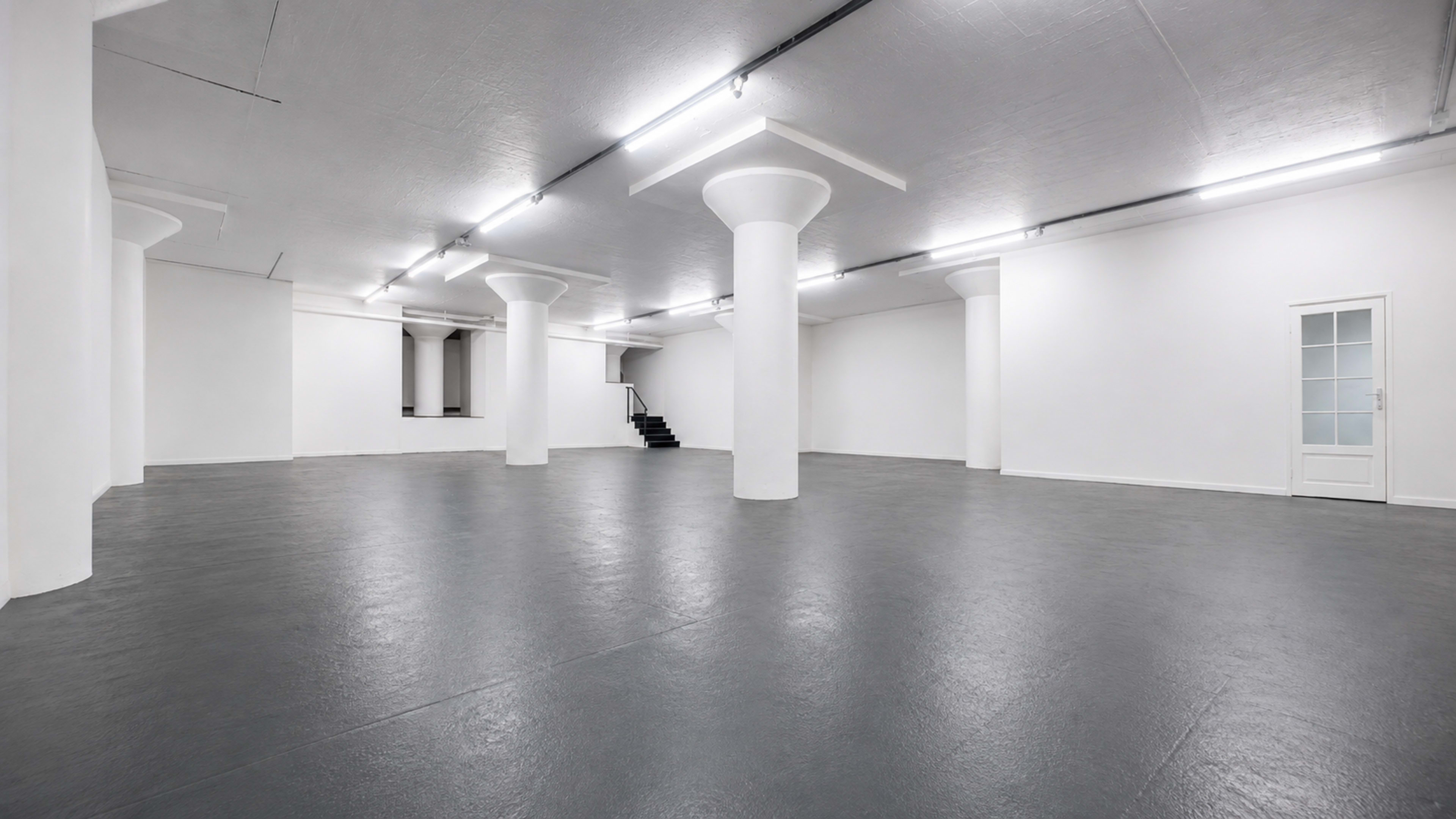 The image shows a spacious, empty room with white walls, gray flooring, and several white columns throughout the area.