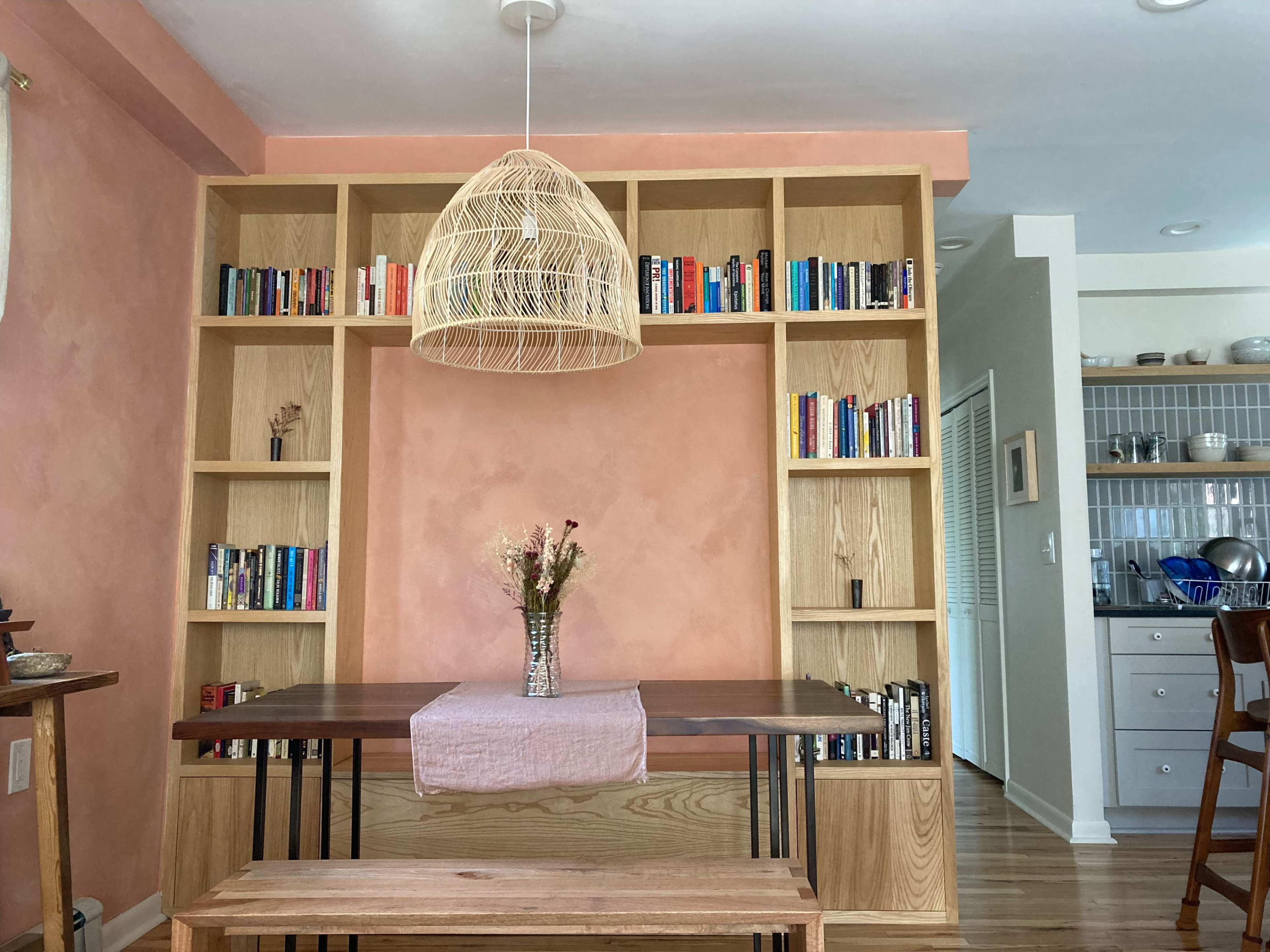 A wooden dining table with a pink table runner, flanked by a bench and a bookshelf filled with colorful books against a peach-colored wall.