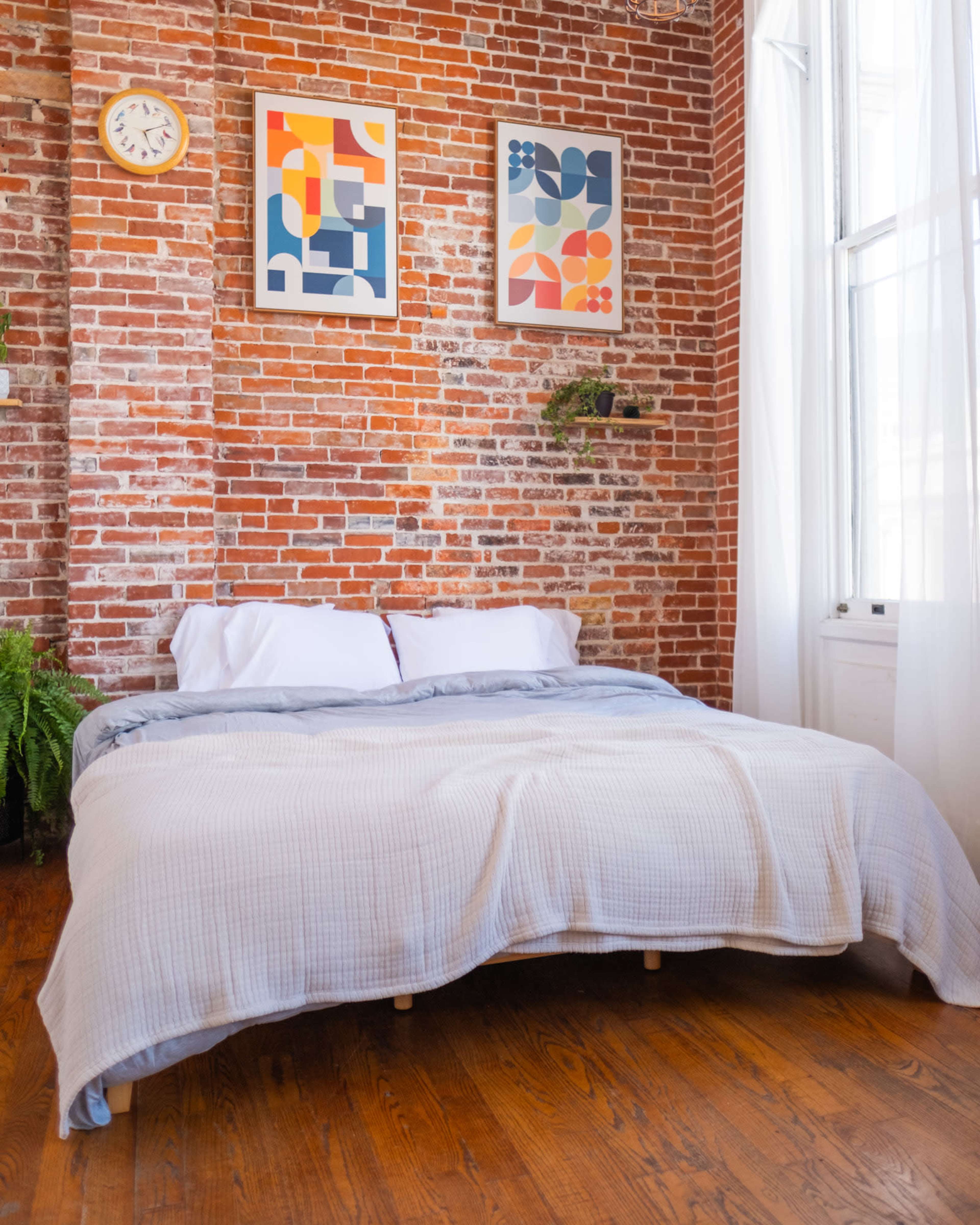 The image shows a cozy bedroom with a wooden floor, a brick wall, a large bed with a light-colored blanket, and two colorful framed artworks above it.