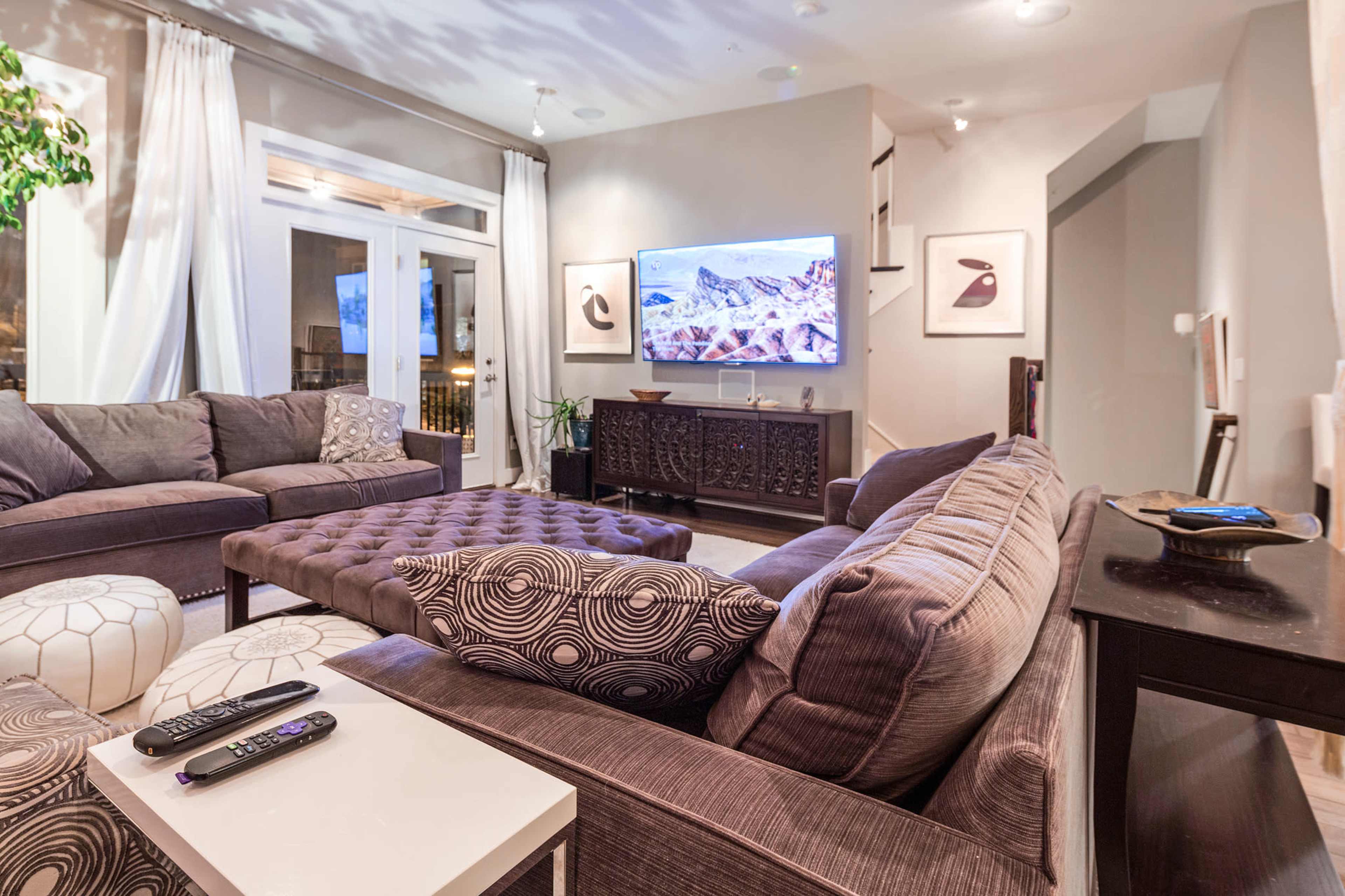 The living room features a large sectional sofa, an ottoman, and a wall-mounted television, with sheer curtains and decorative items enhancing the space.