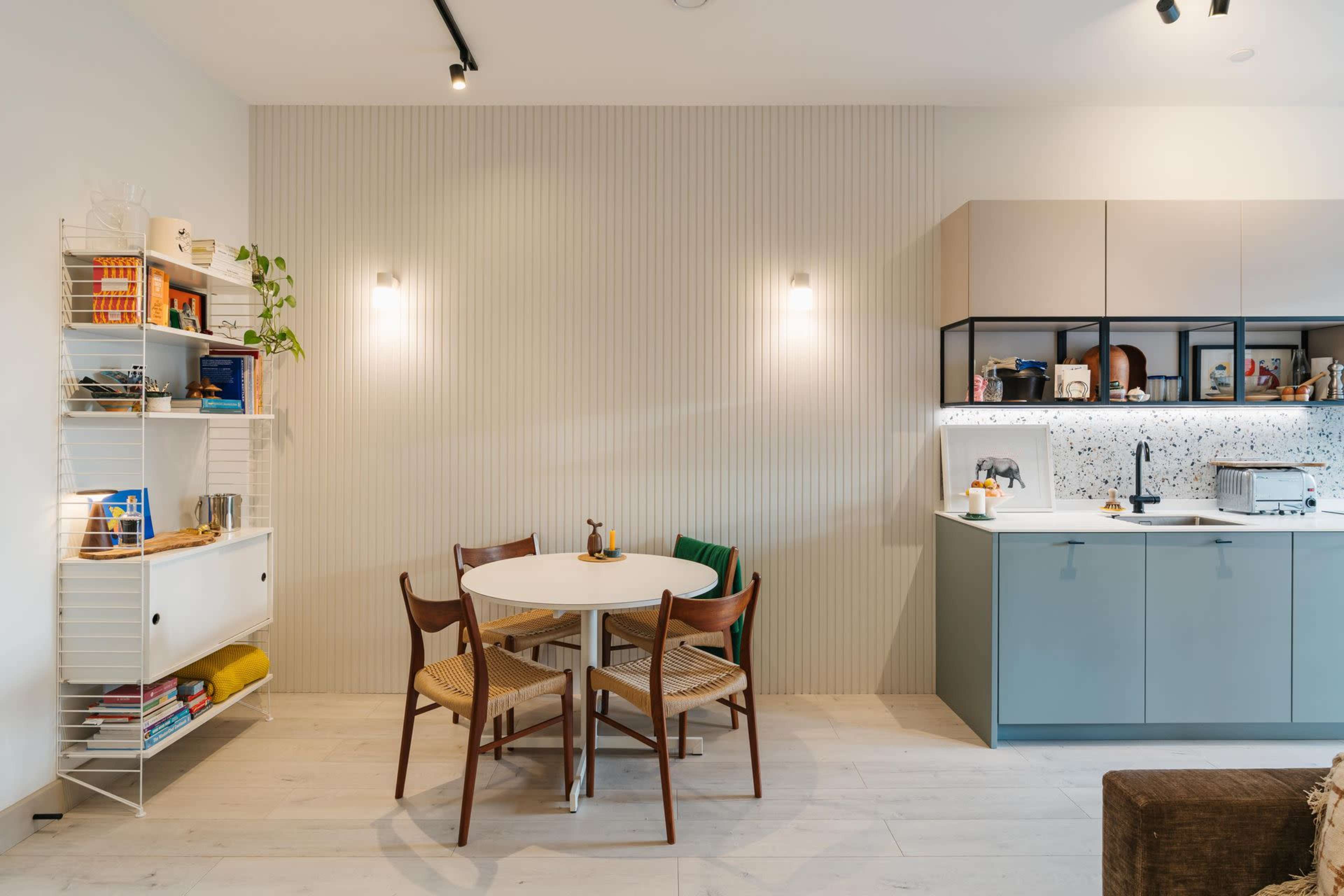 The image shows a modern kitchen and dining area featuring a round table with four chairs, a minimalist kitchen with cabinets, and a white shelving unit filled with books and plants.