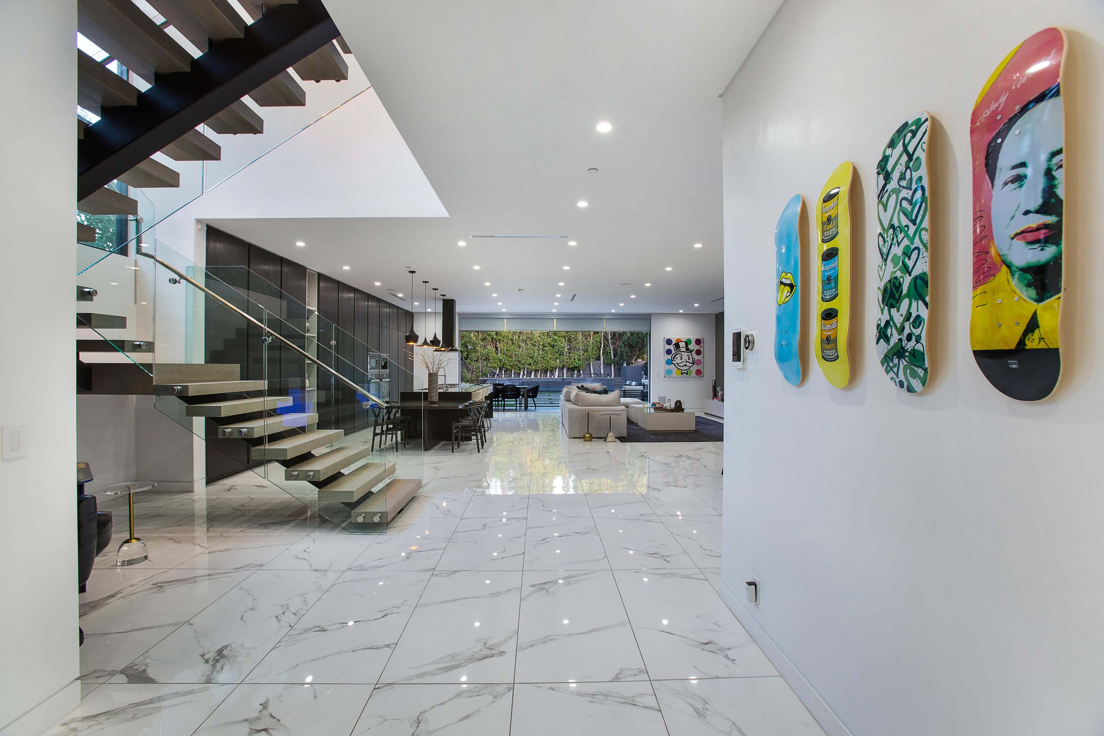 The image shows a modern, open-concept living space with marble flooring, a staircase, and colorful skateboard art on the wall.