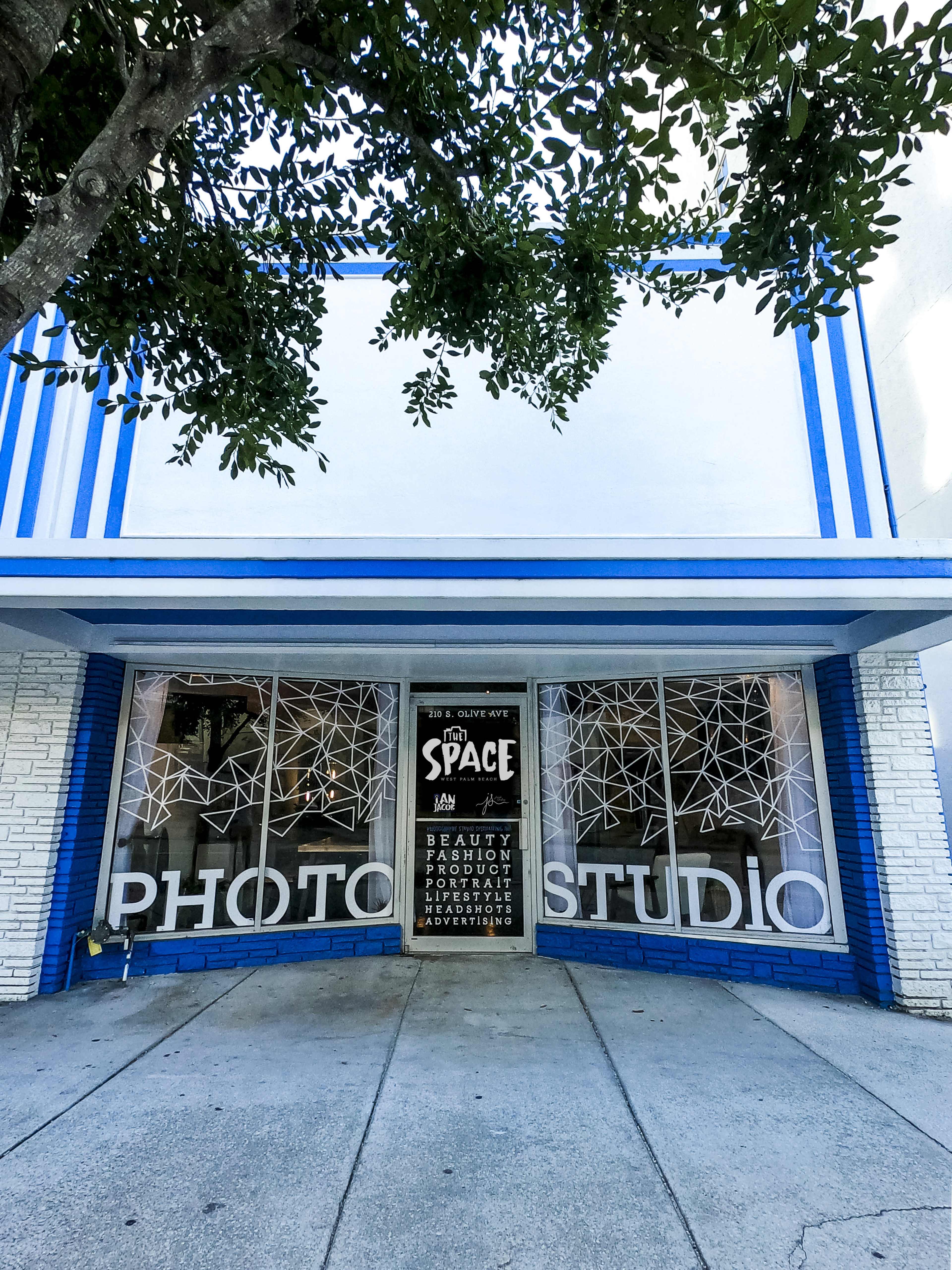 A photo studio with large windows displaying geometric designs and a sign advertising beauty, fashion, and adventure is located beneath a blue and white building facade.