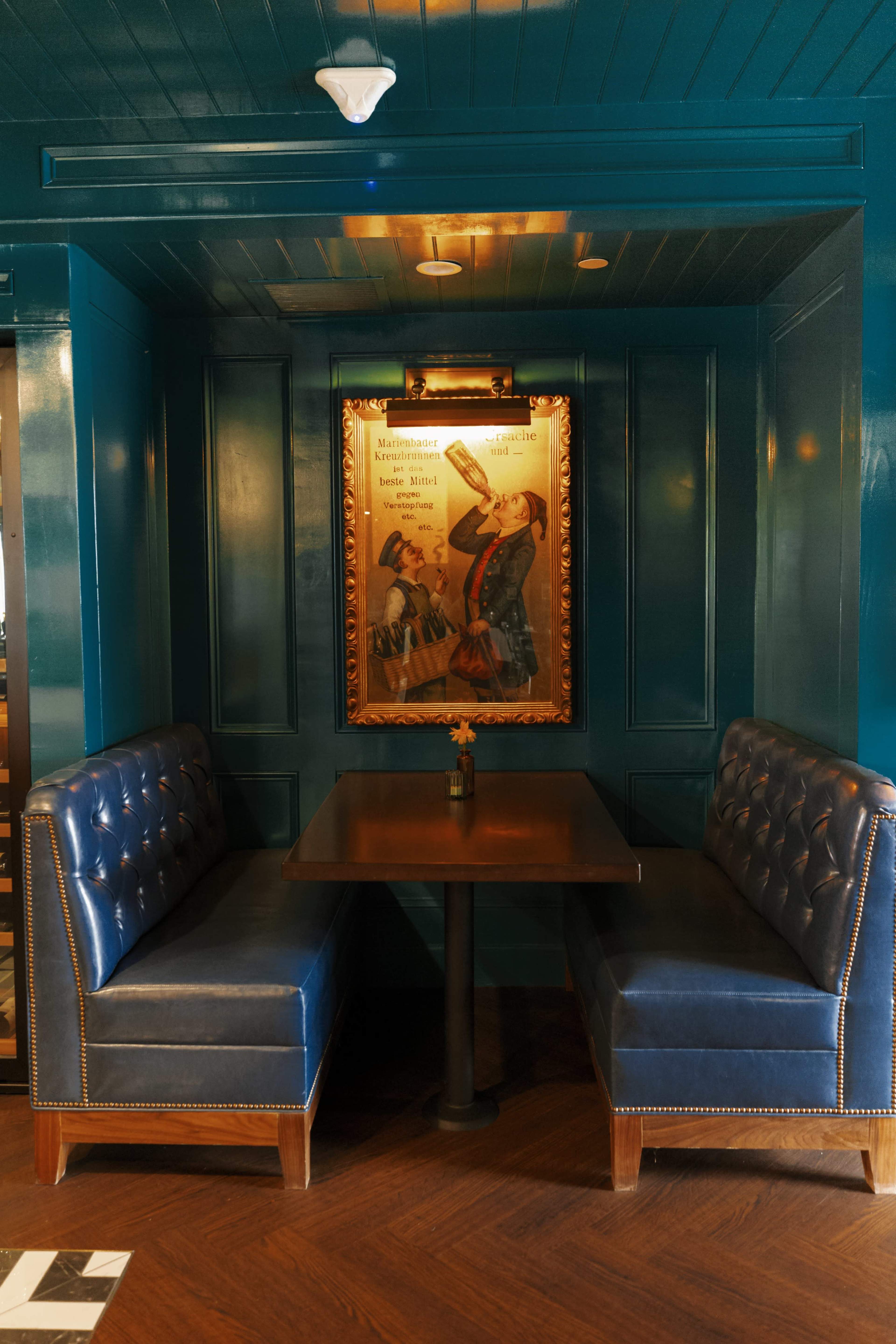 A cozy dining nook features two blue upholstered booths flanking a dark wood table beneath a framed vintage artwork on a deep green wall.