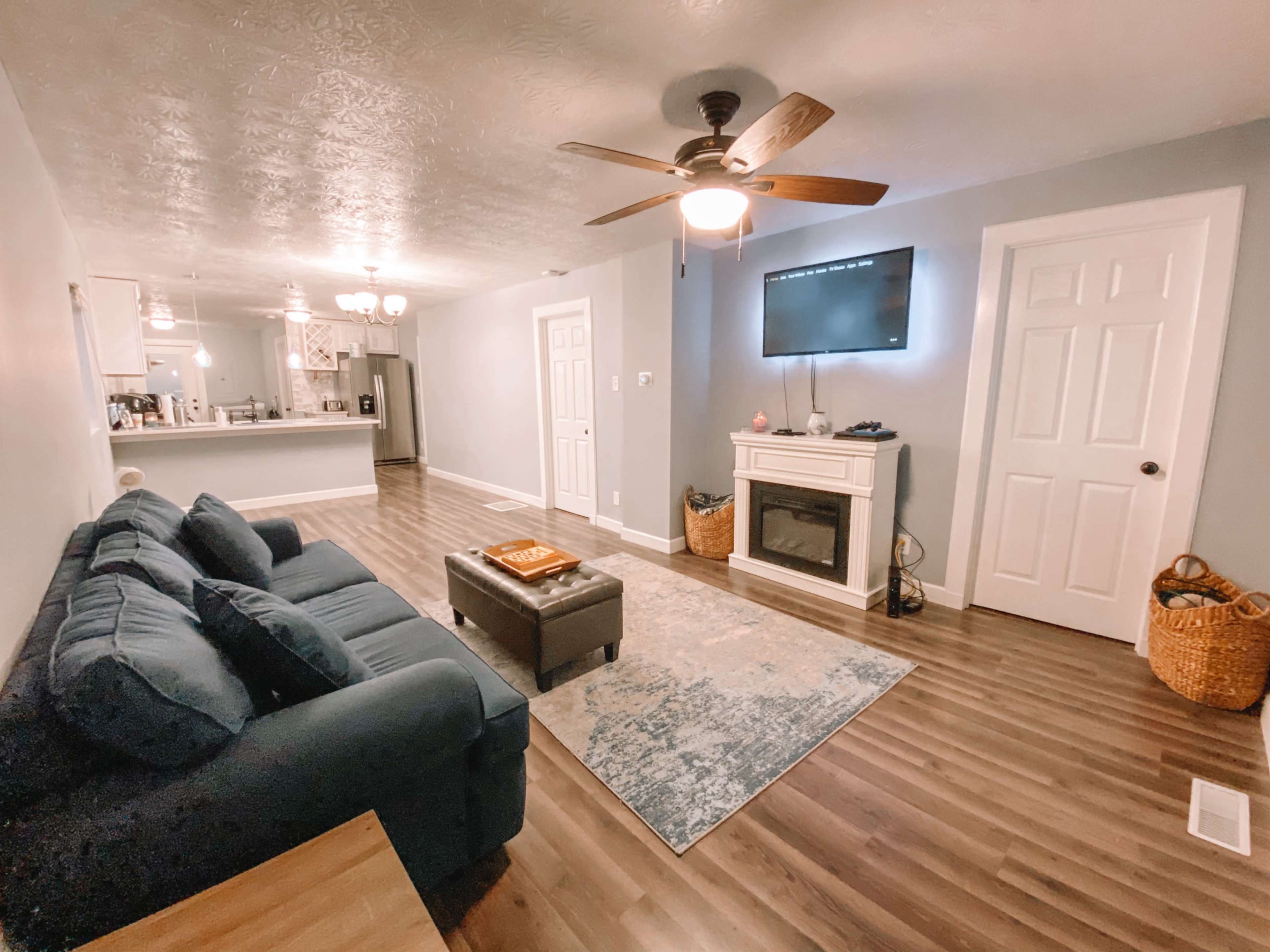 The living room features a blue sofa facing a wall-mounted television above a fireplace, with a coffee table and a patterned rug on the wooden floor.