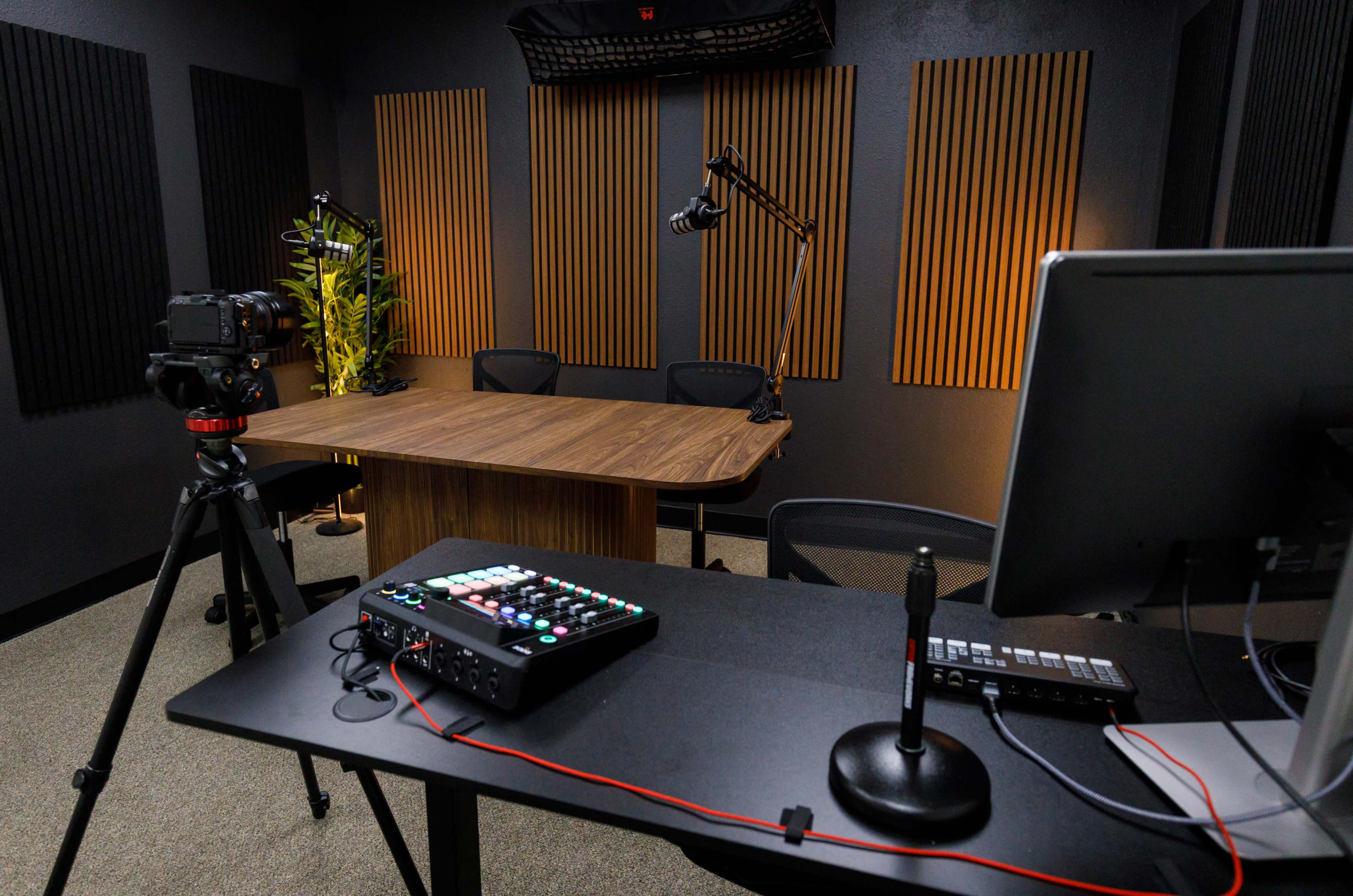 The image shows a recording studio with a wooden table, two chairs, and audio equipment, including a camera on a tripod and a control panel on a desk.