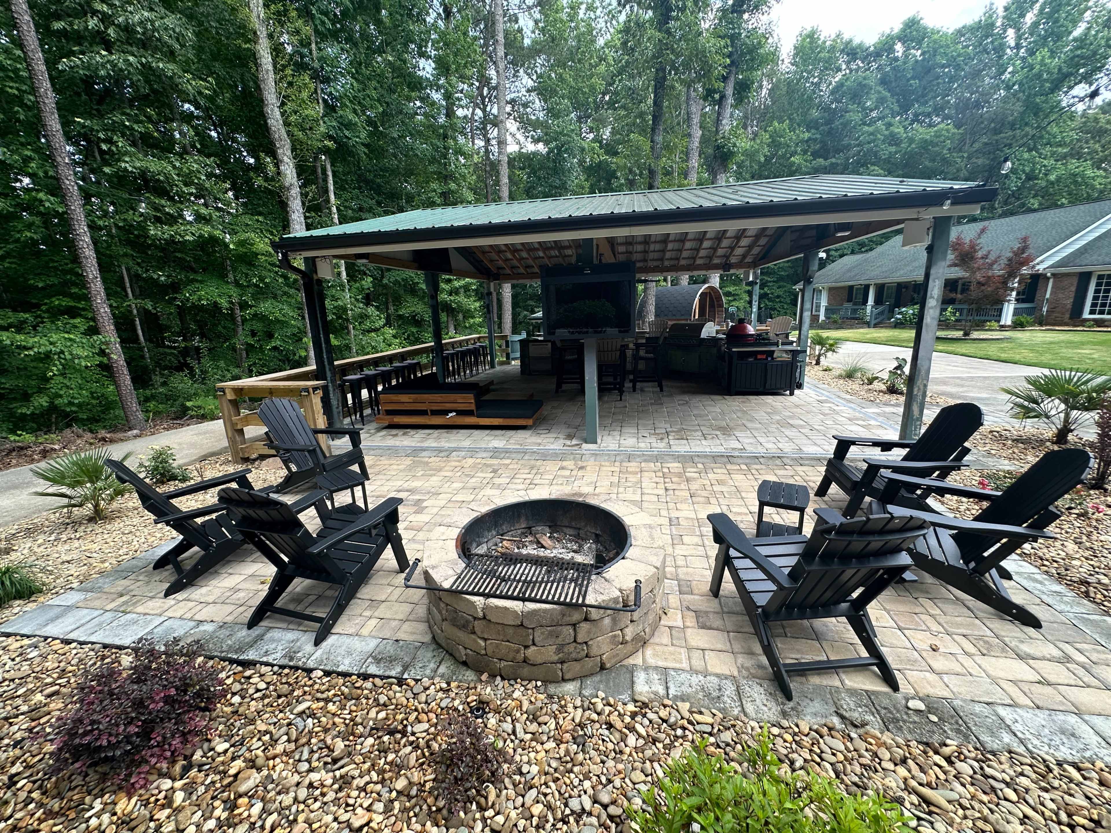 A covered outdoor area features a stone patio with a fire pit surrounded by black chairs and a grill, set in a wooded backdrop.
