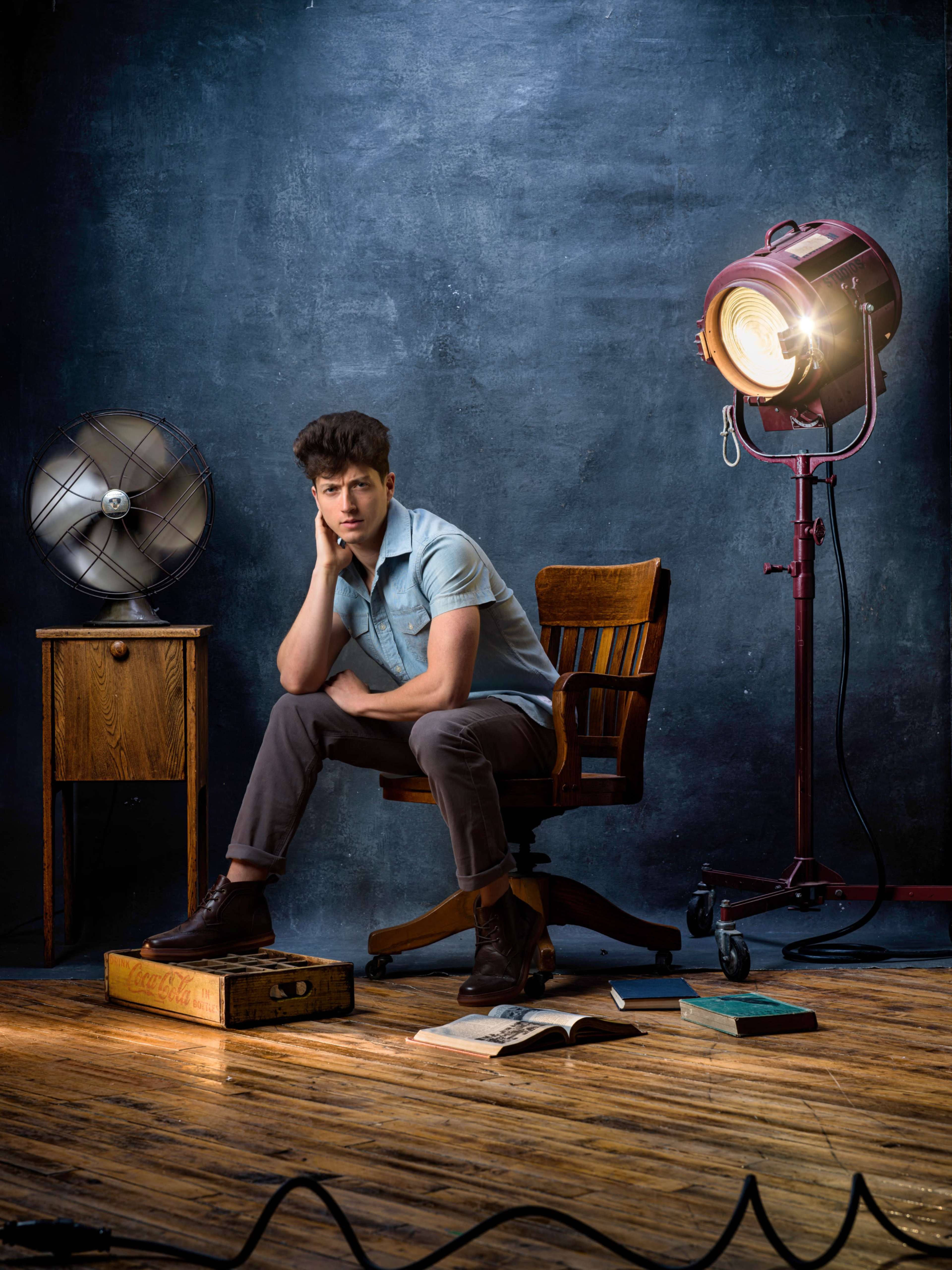 A young man sits thoughtfully on a wooden chair in a dimly lit studio, surrounded by a spotlight, a fan, and scattered books on the floor.