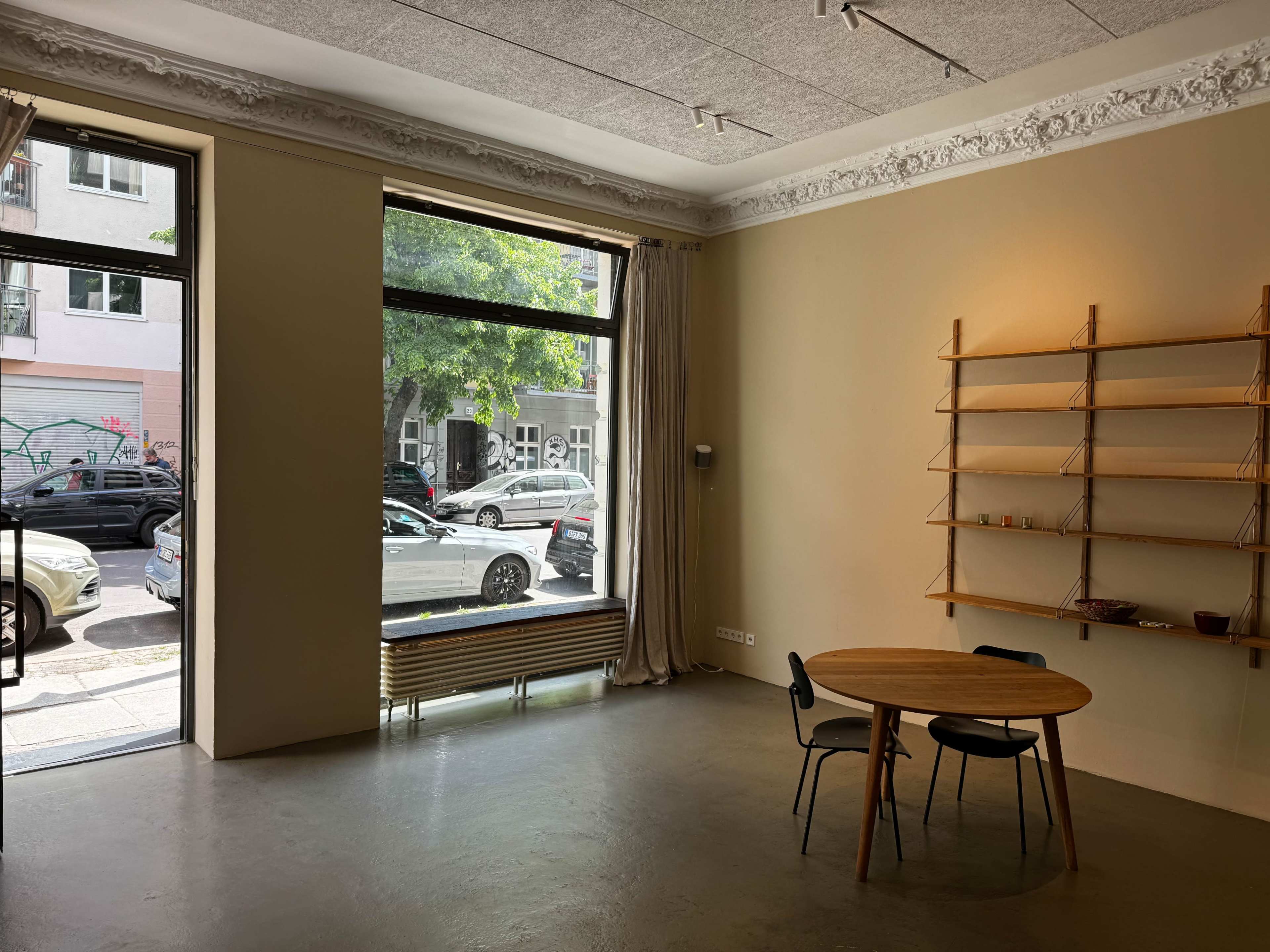 The image shows a minimalist interior space with a round wooden table and two chairs, featuring large windows that open to a view of parked cars and greenery outside.