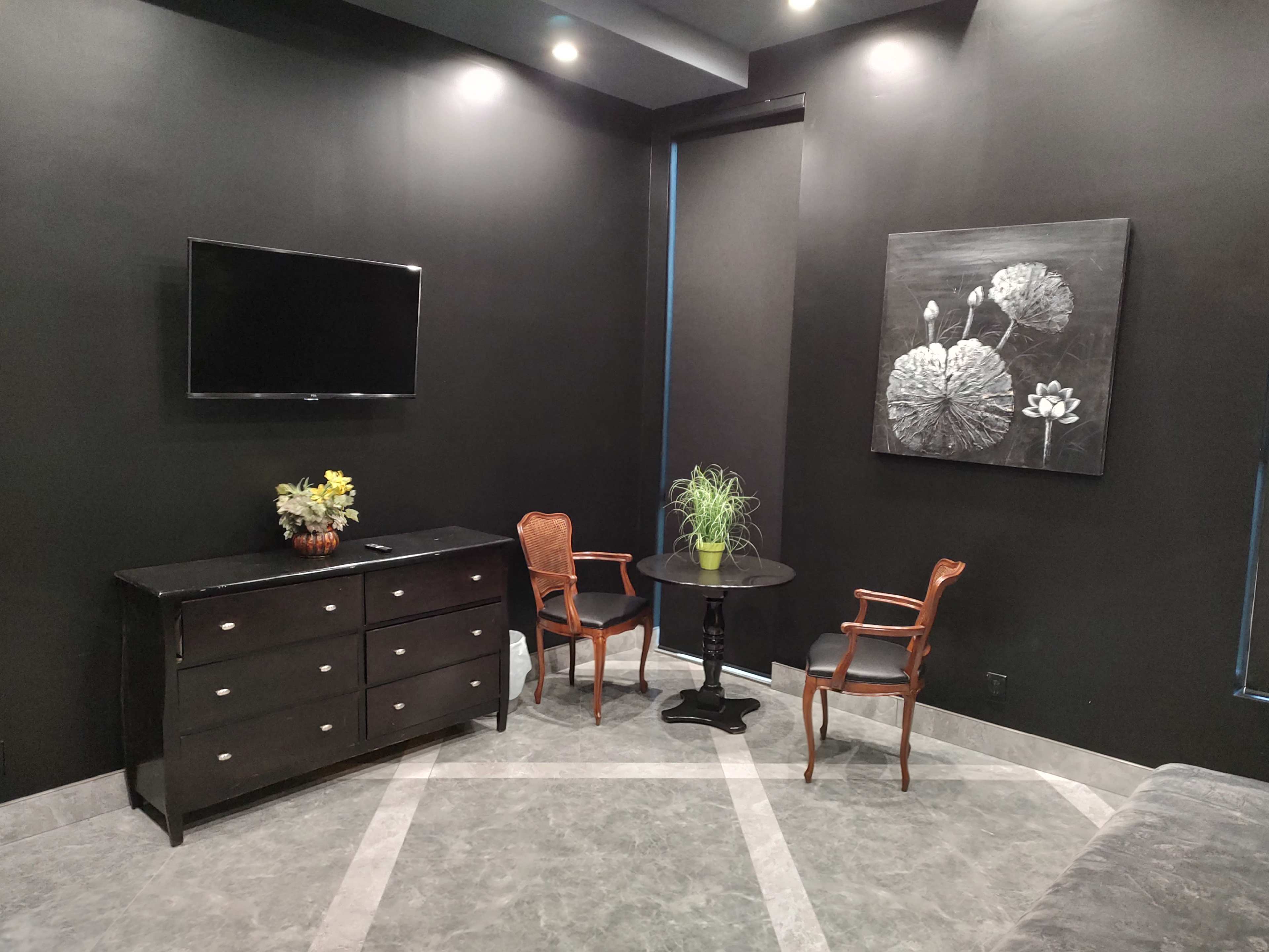 A small, modern room with black walls, a television mounted on the wall, a dresser, a round table with two chairs, and a decorative plant.