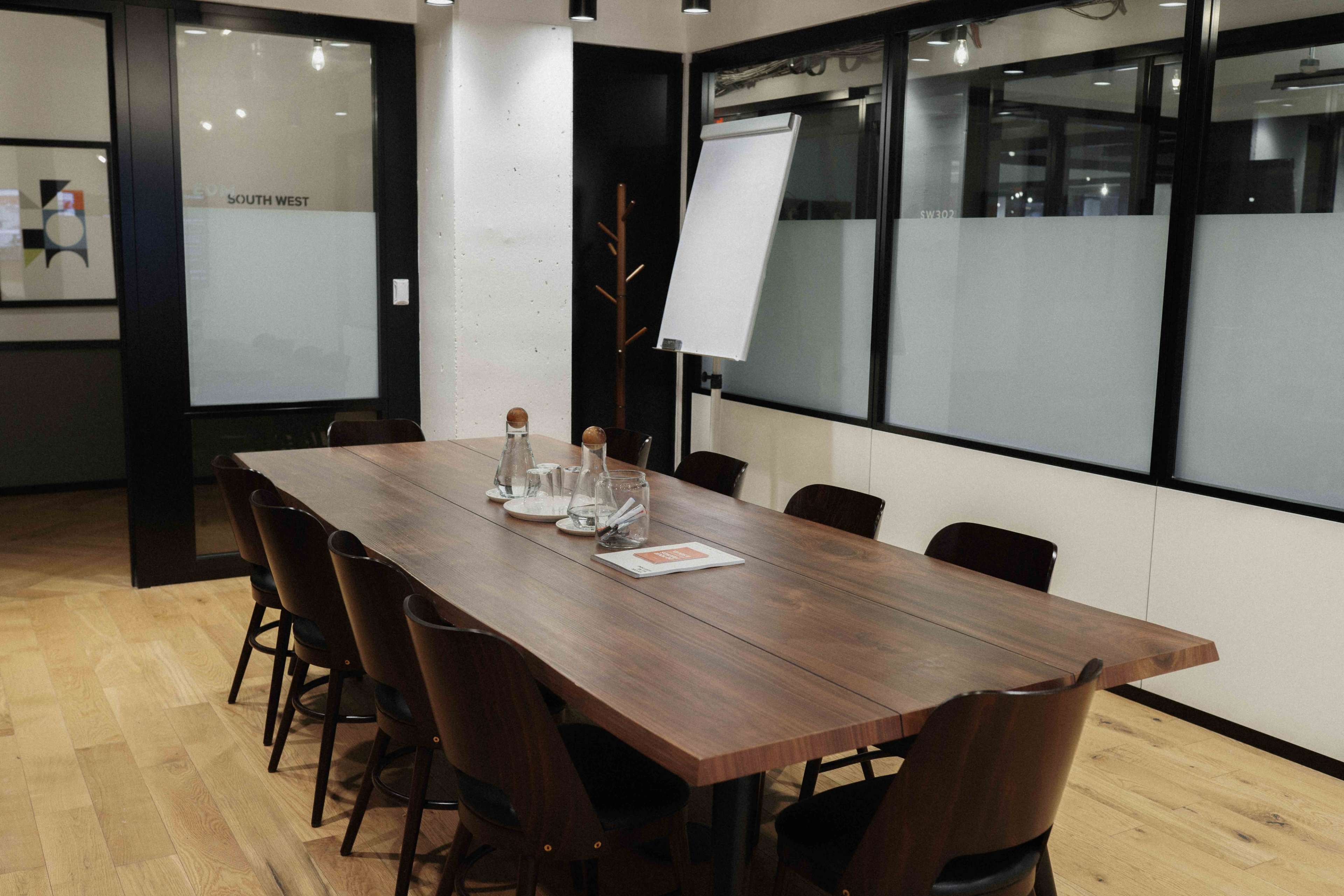 A long wooden conference table with chairs is set up in a modern meeting room, accompanied by water pitchers and a notepad.