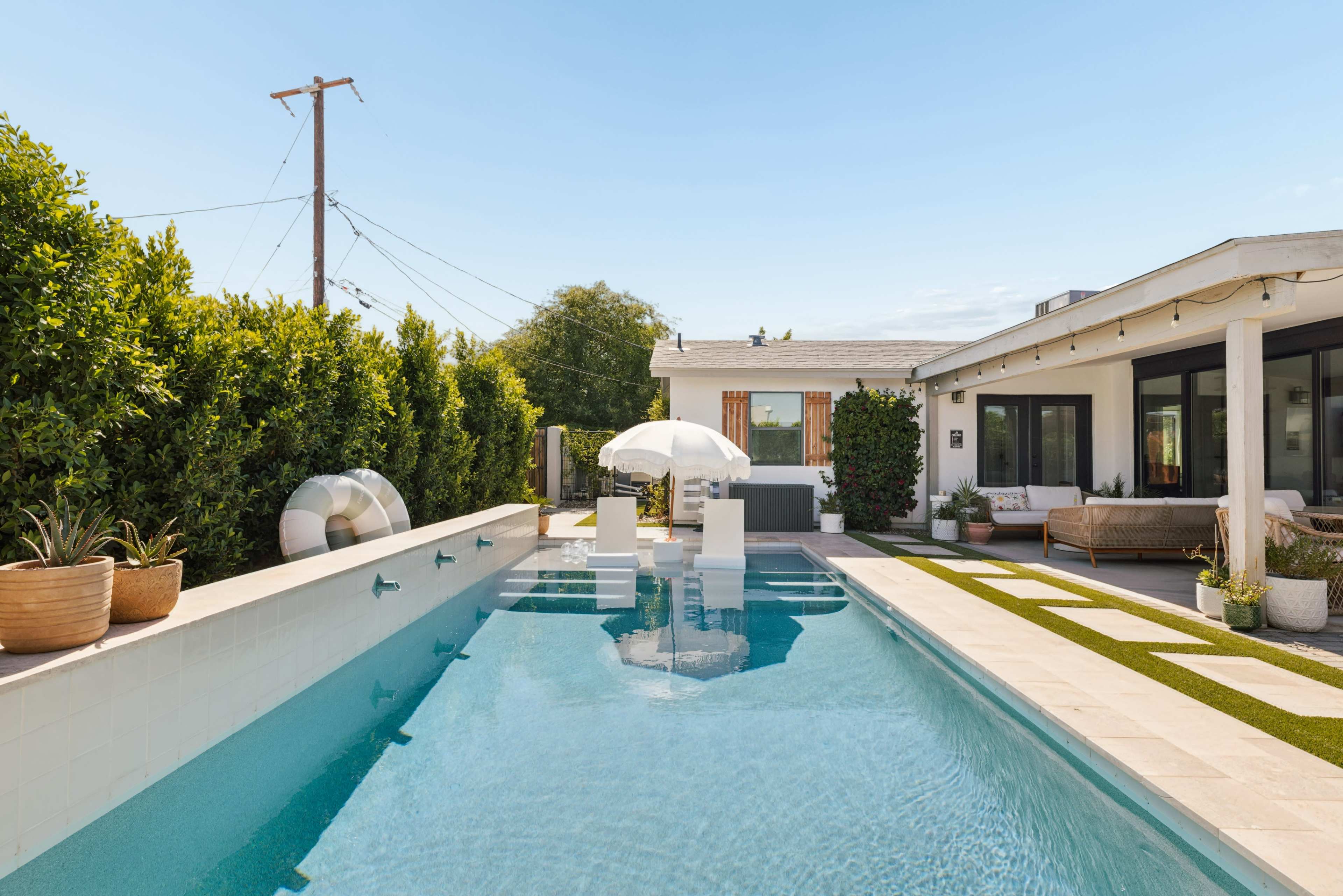 The image shows a modern backyard with a swimming pool, lounge area, and neatly trimmed hedges under a clear sky.