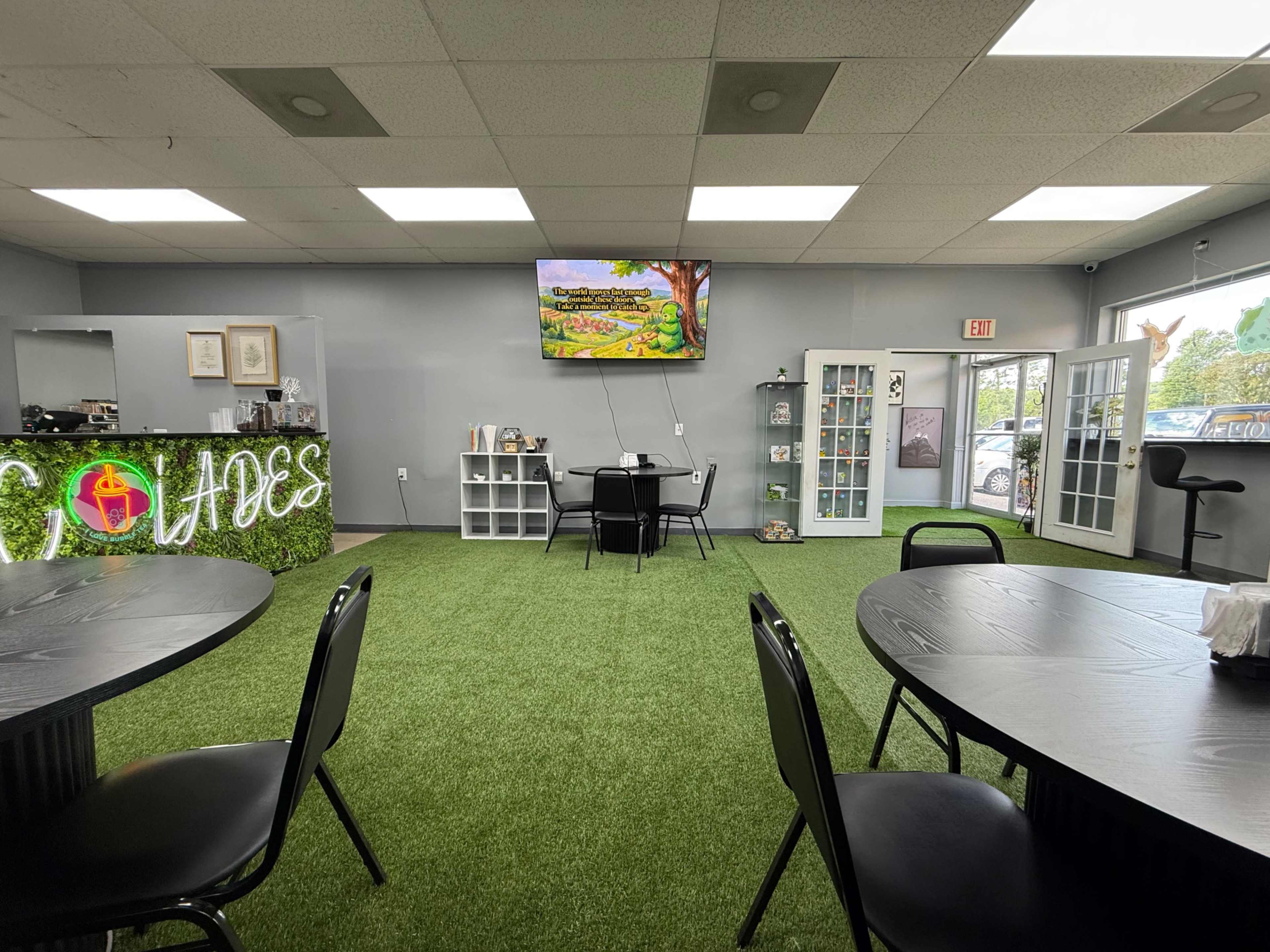 The image shows an indoor space featuring several black tables on a green artificial grass floor, a wall with the word "Clouds" illuminated, and a television displaying a landscape scene.
