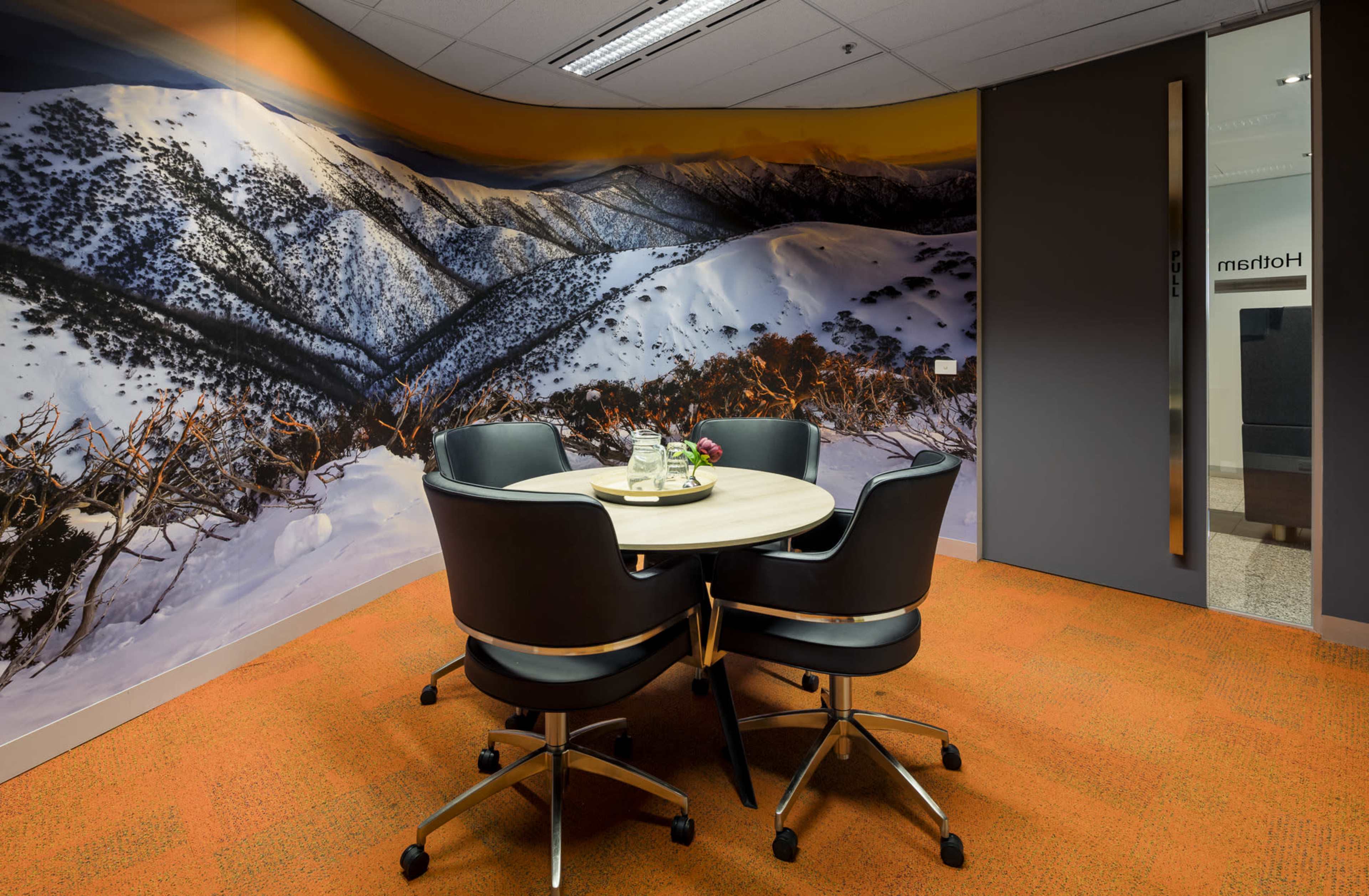 The image shows a meeting room with a round table and four chairs, featuring a large mountain landscape mural on one wall and orange carpet flooring.