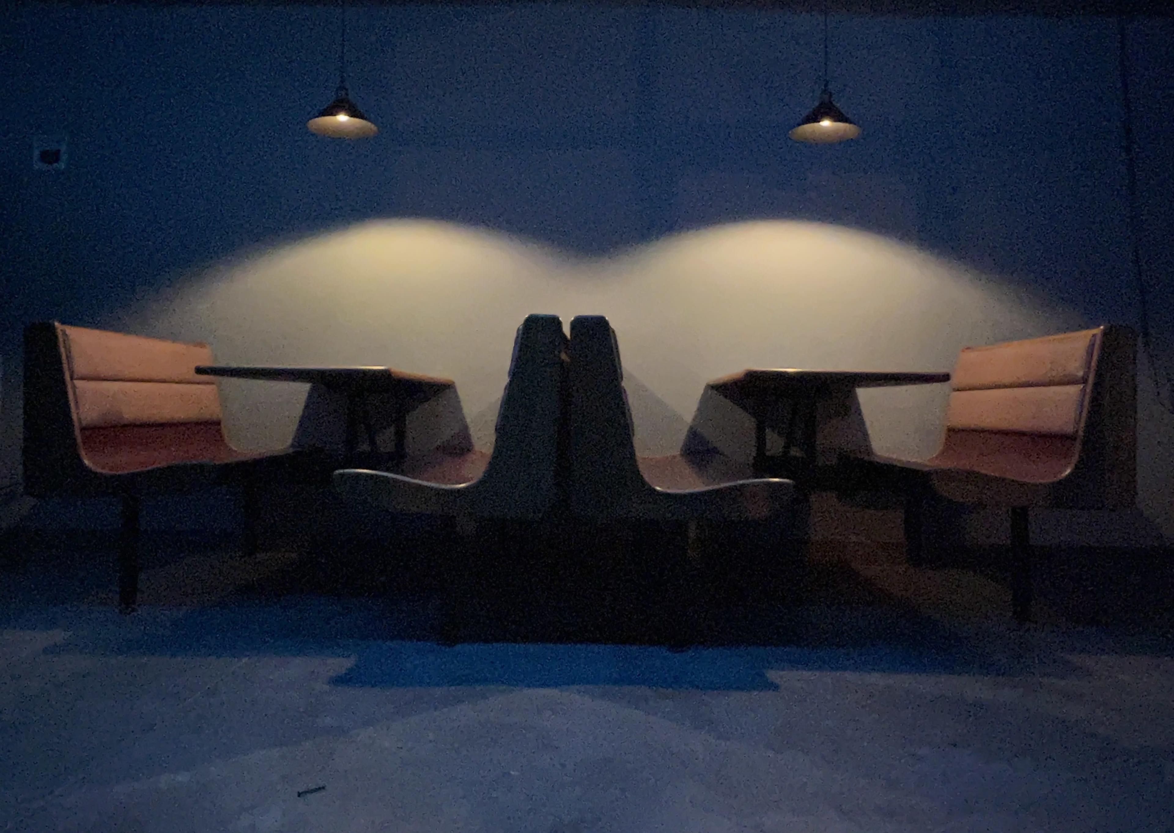 The image shows a dimly lit interior with two sets of booth seating and overhead lights casting shadows on the walls.