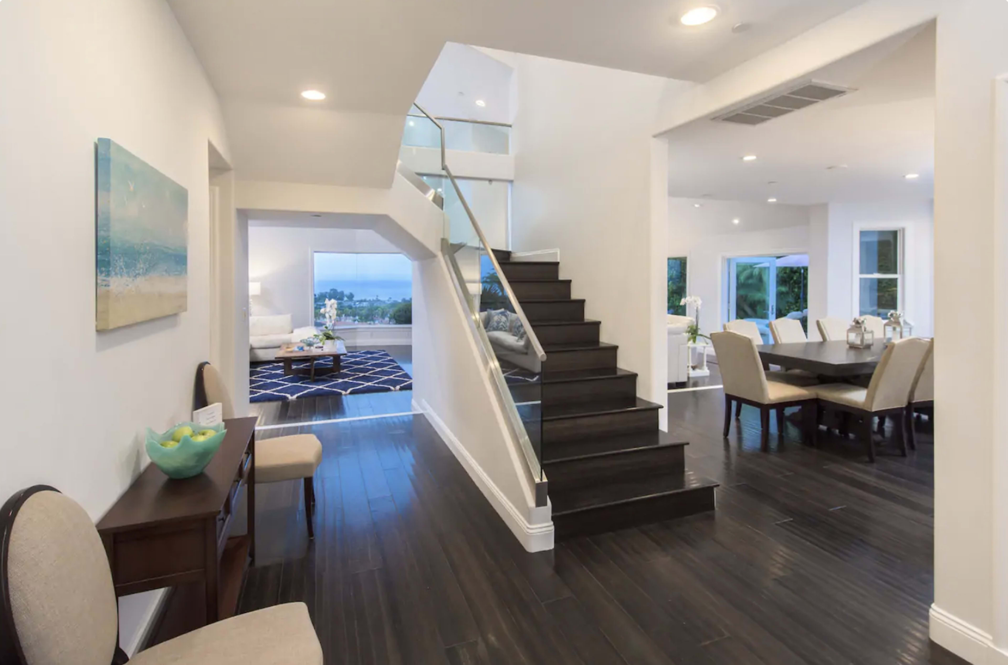 A modern entryway with a staircase leading to an upper level, adjacent to a dining area and a living room with large windows.