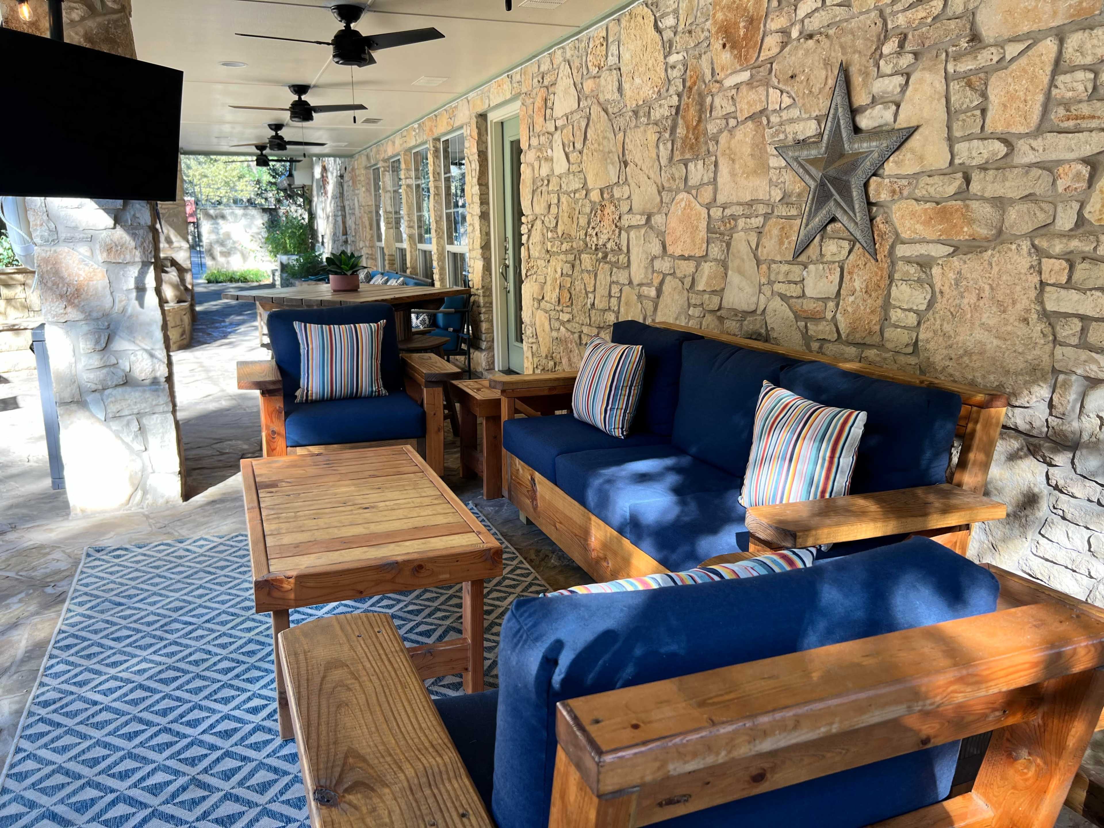 The image shows a comfortable outdoor seating area with blue sofas, wooden tables, and stone walls, adorned with decorative items like pillows and a star.