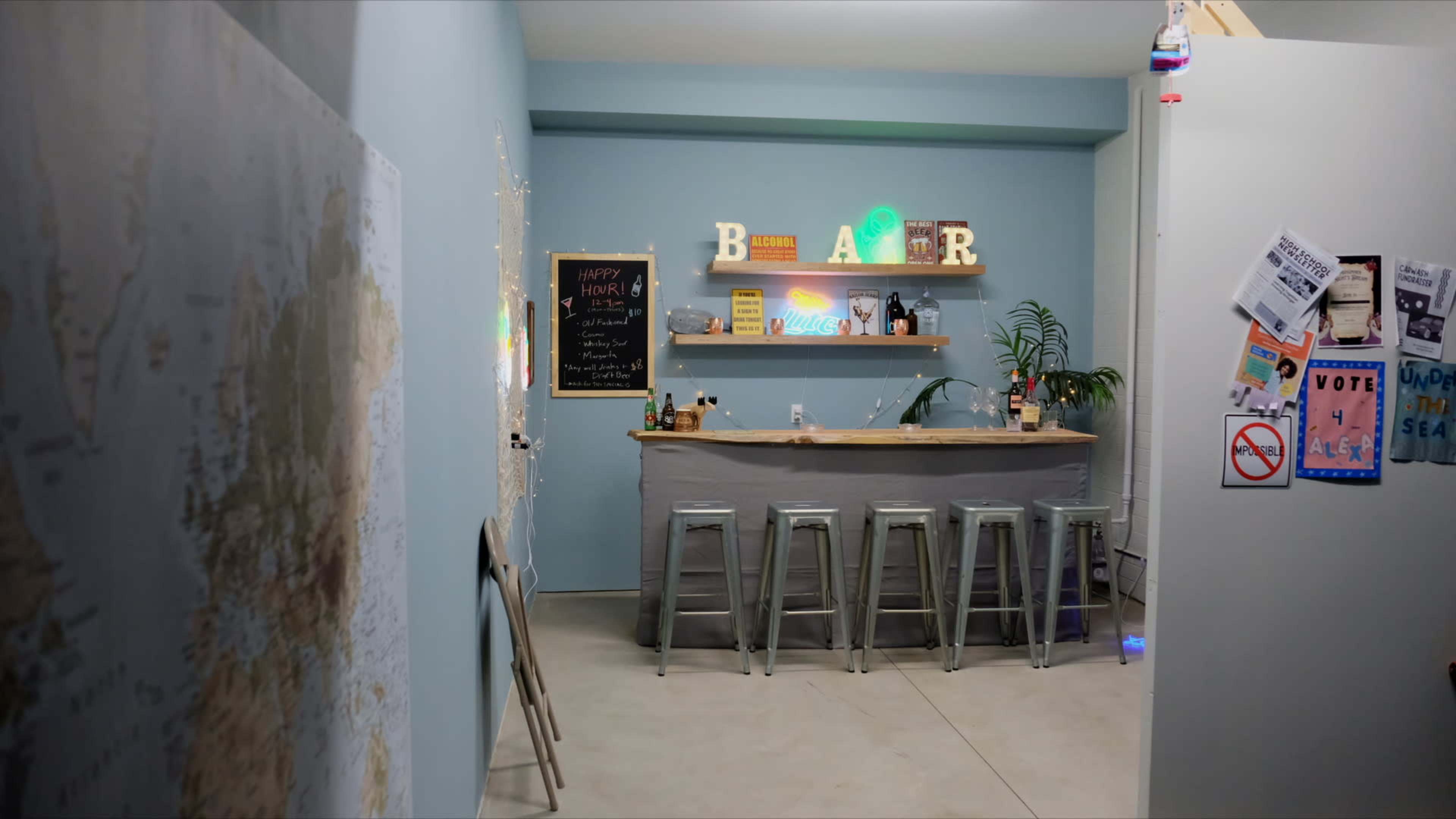 The image shows a bar area with a countertop, high stools, colorful neon lights, and a chalkboard menu in a light blue room.