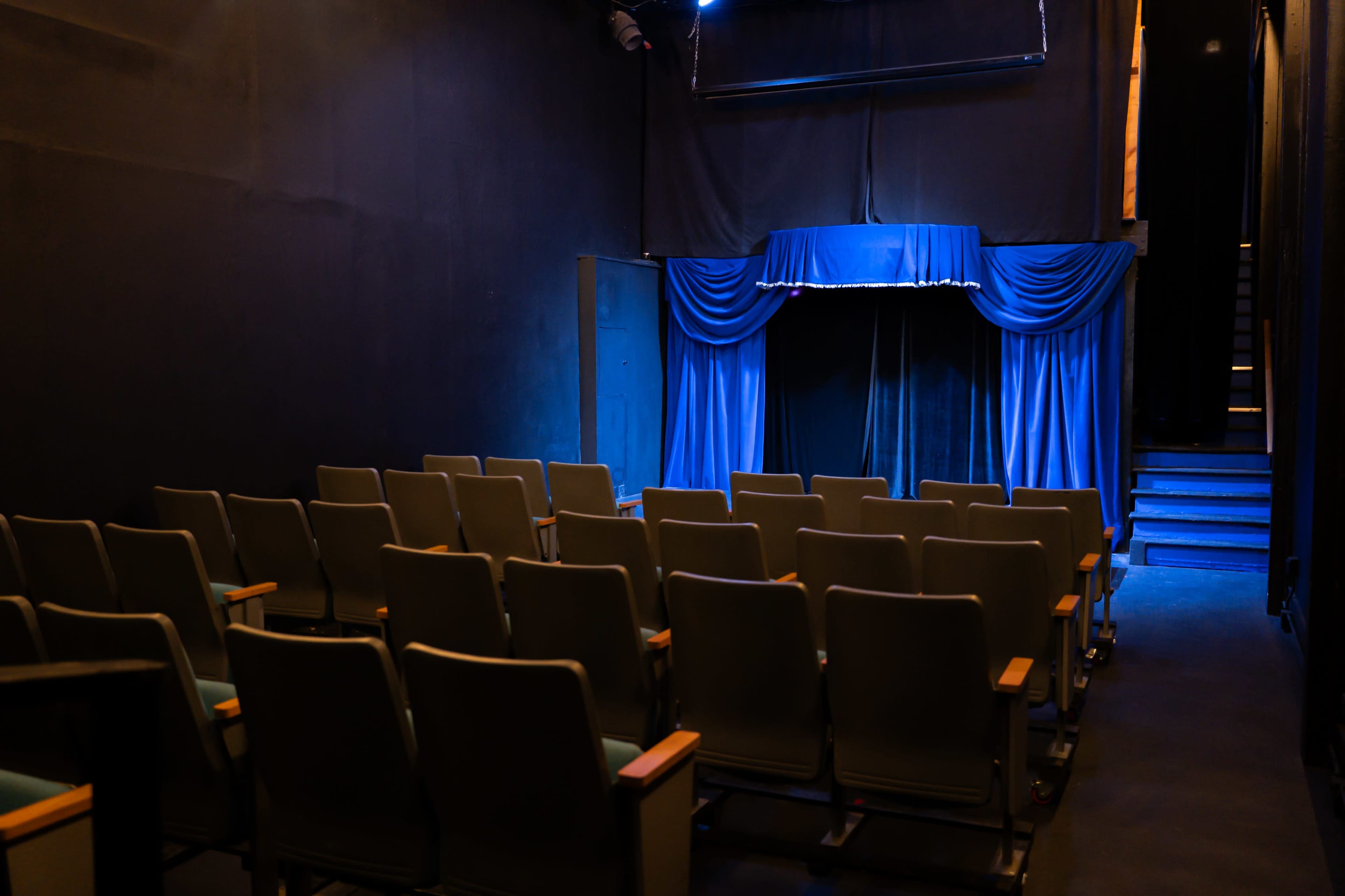 The image shows a dimly lit theater with empty rows of gray seats facing a small stage adorned with blue curtains.