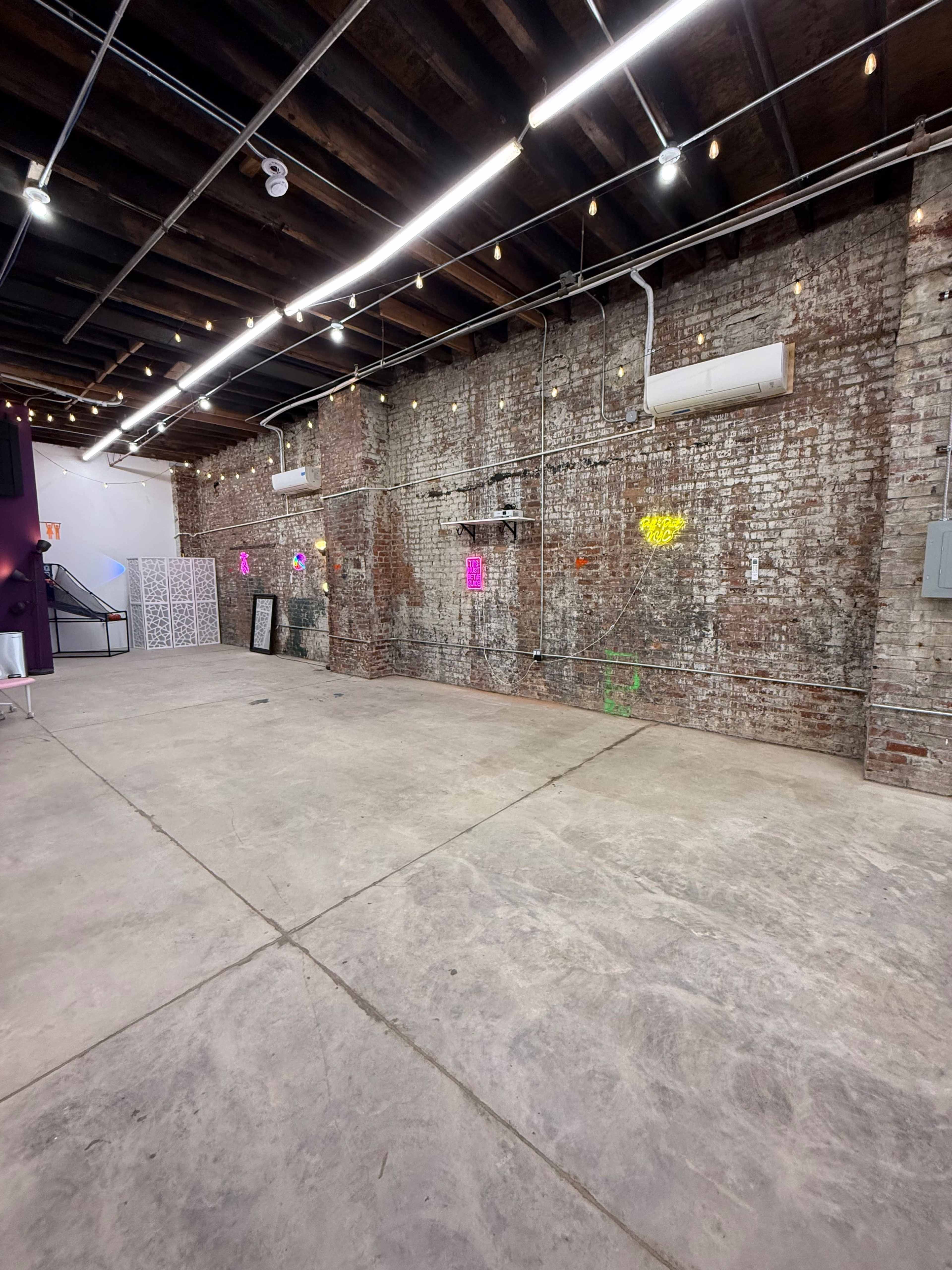 The image shows an empty warehouse space with exposed brick walls, concrete flooring, and several colorful neon lights on the wall.