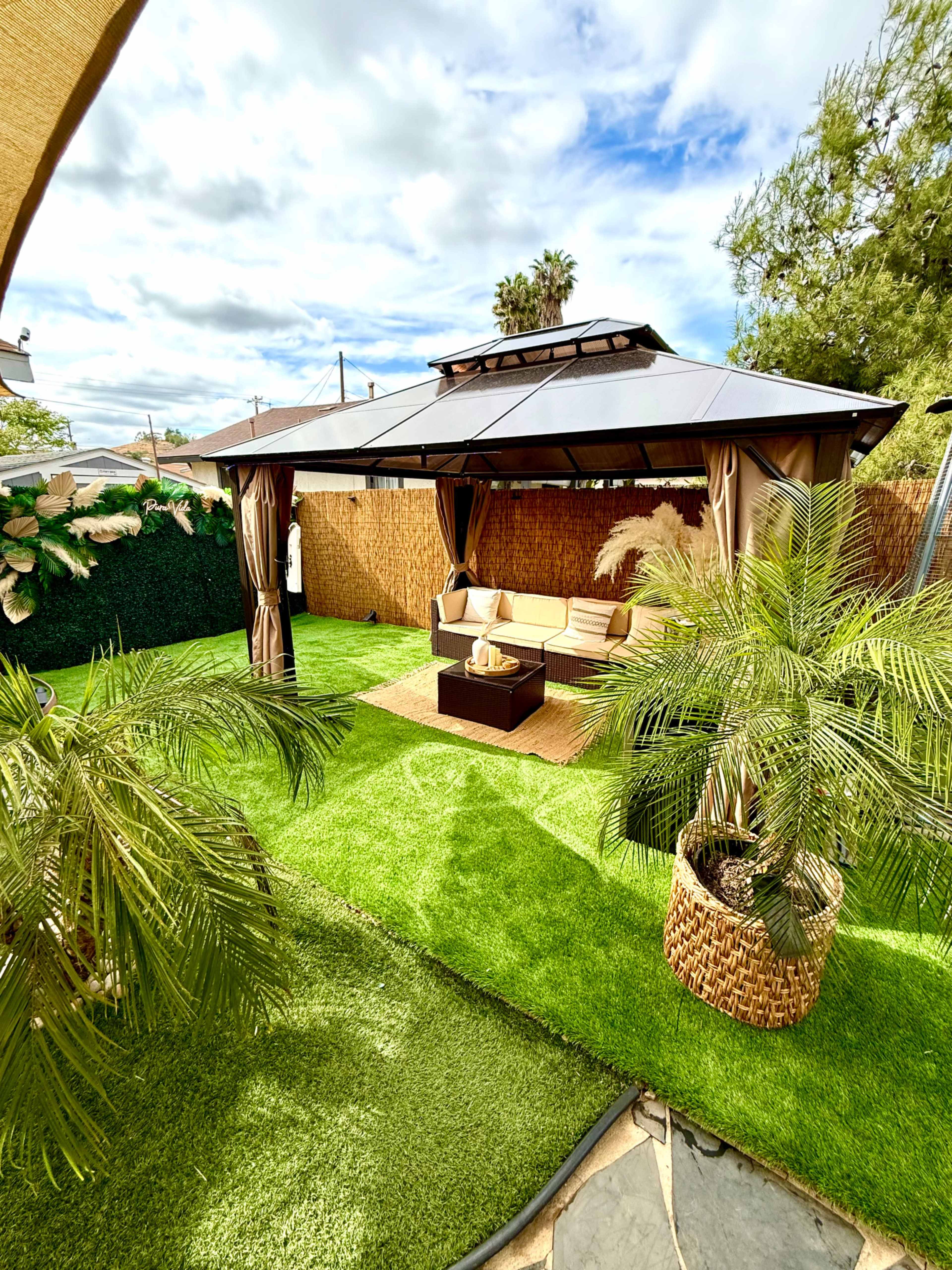 The image shows a well-maintained outdoor gazebo with a seating area on a grassy lawn, surrounded by palm plants and a privacy fence.