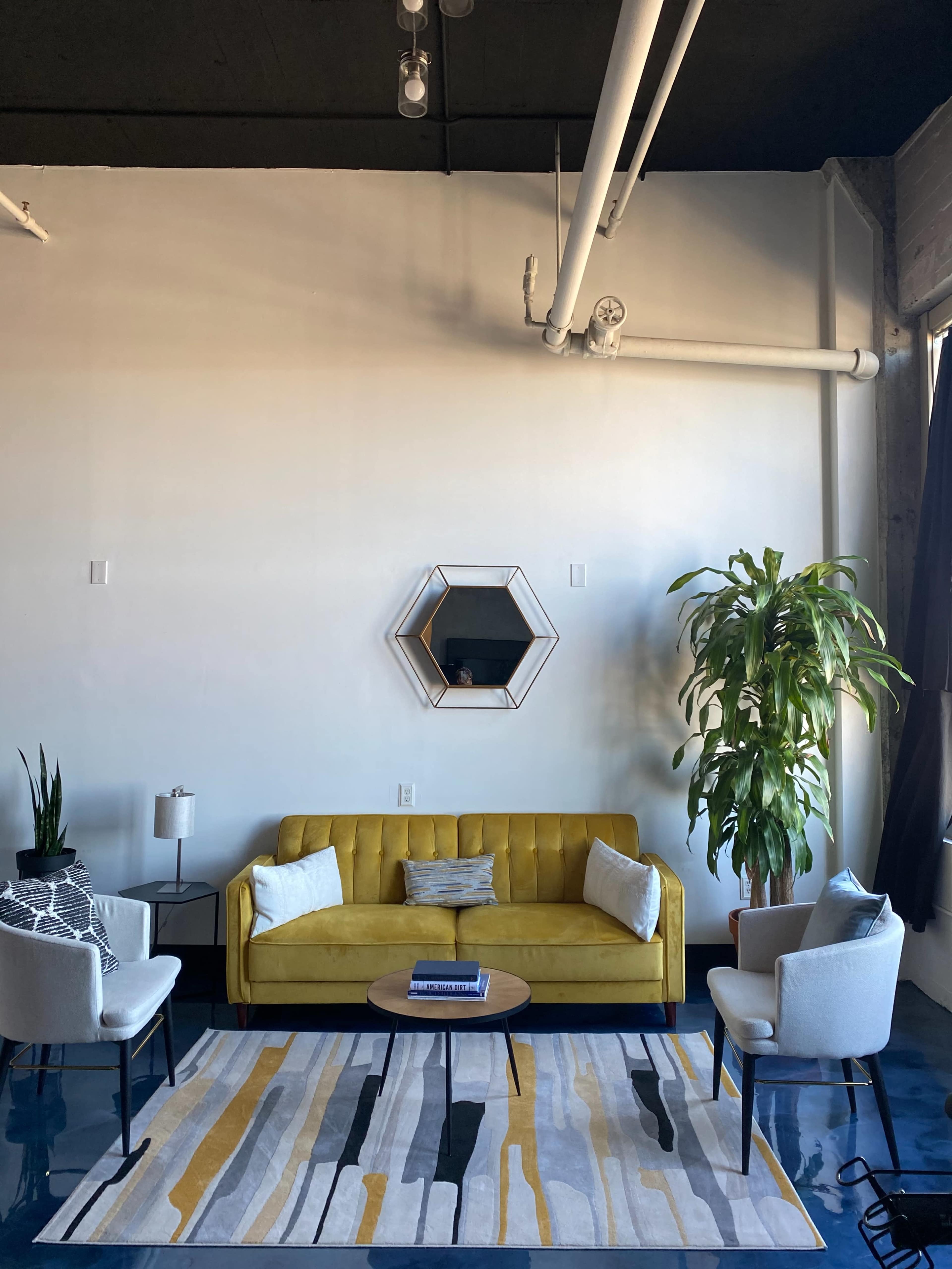 A modern living room with a yellow sofa, two light-colored chairs, a round coffee table, a large plant, and a geometric mirror on the wall.