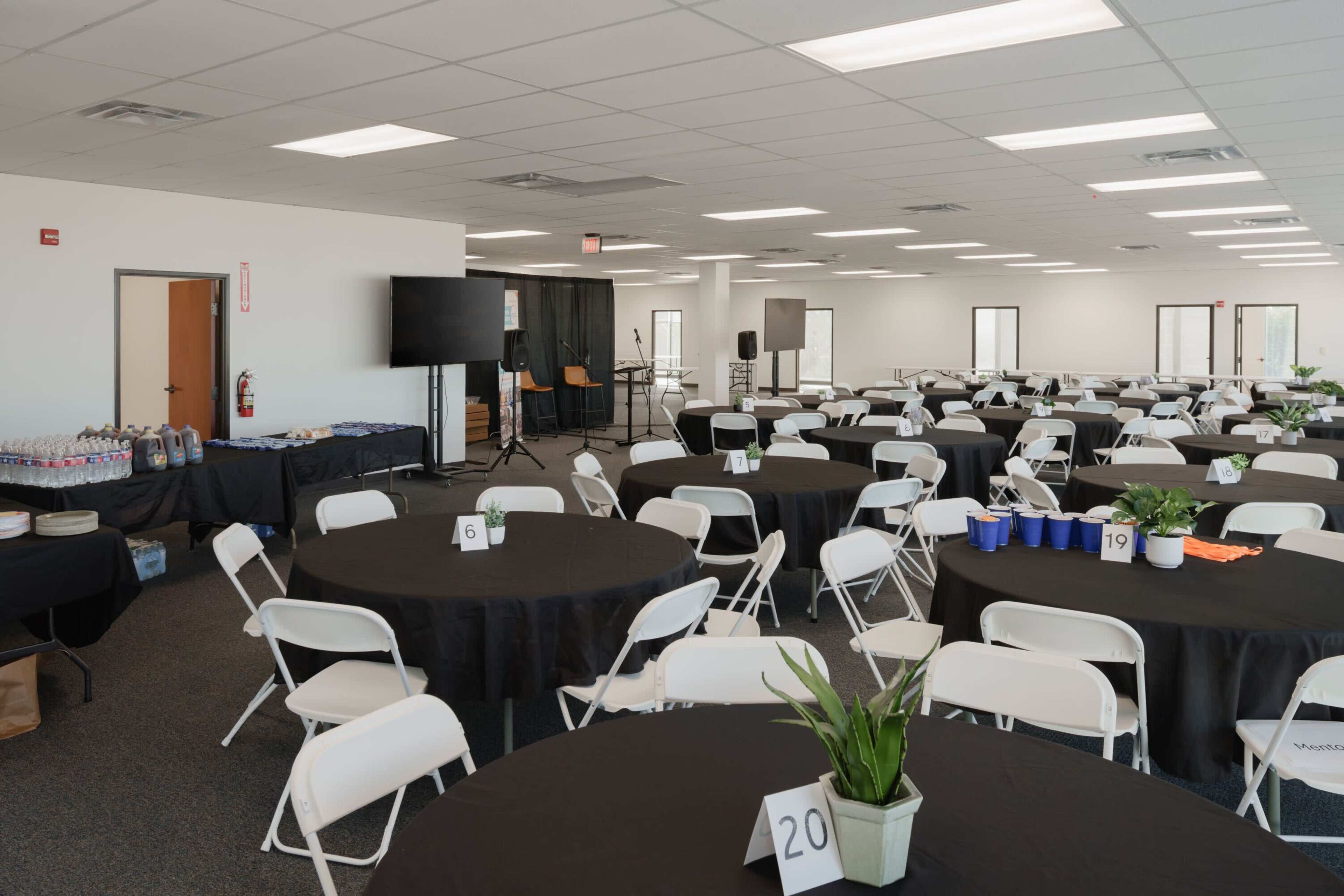 The image shows a large, empty event space with several round tables set up, each with a black tablecloth, and equipment positioned for presentations in the background.