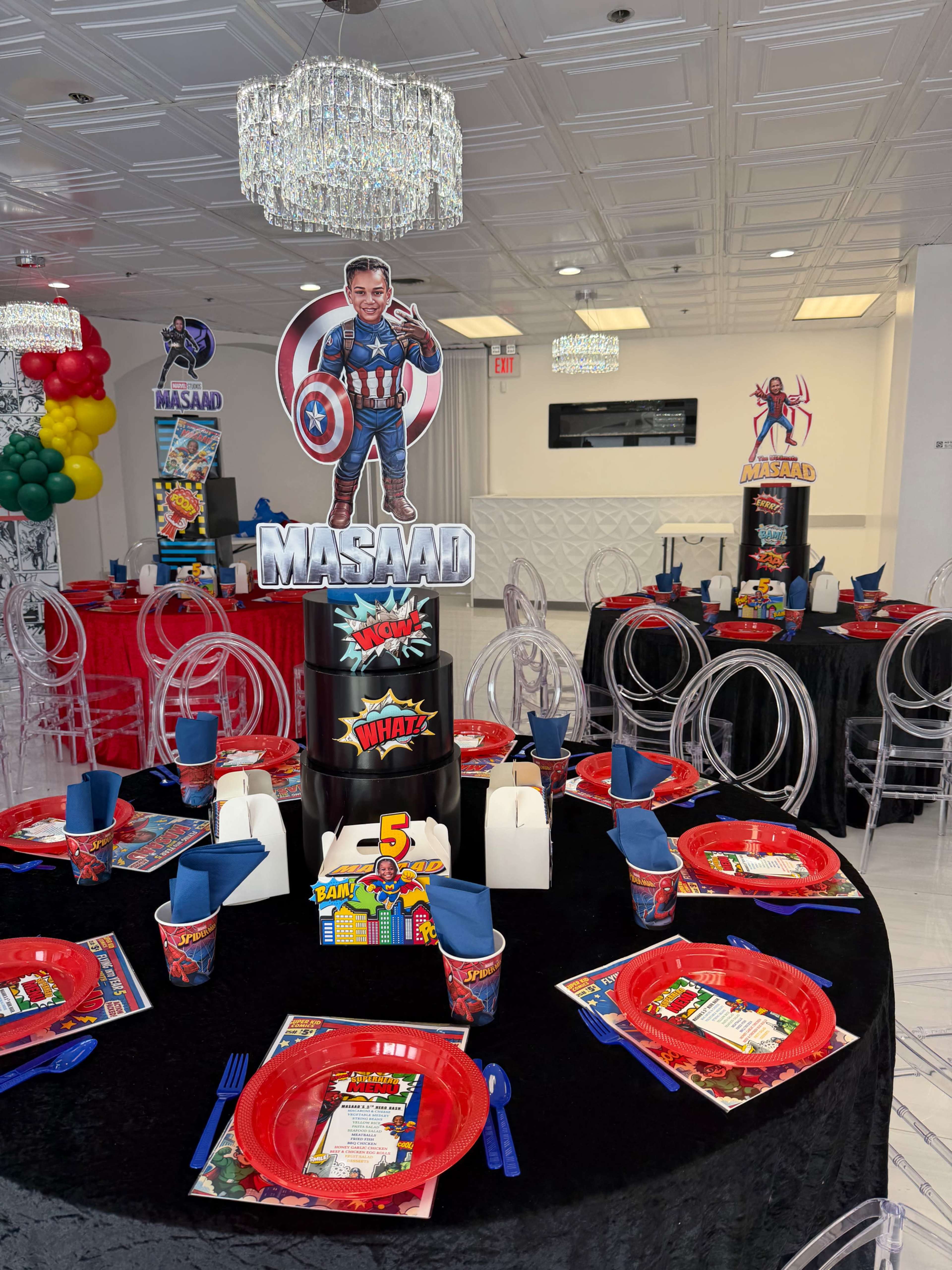 A children's birthday party setup features tables decorated with red and black tablecloths, party-themed centerpieces, and superhero cutouts, along with plates and cups displaying colorful designs.