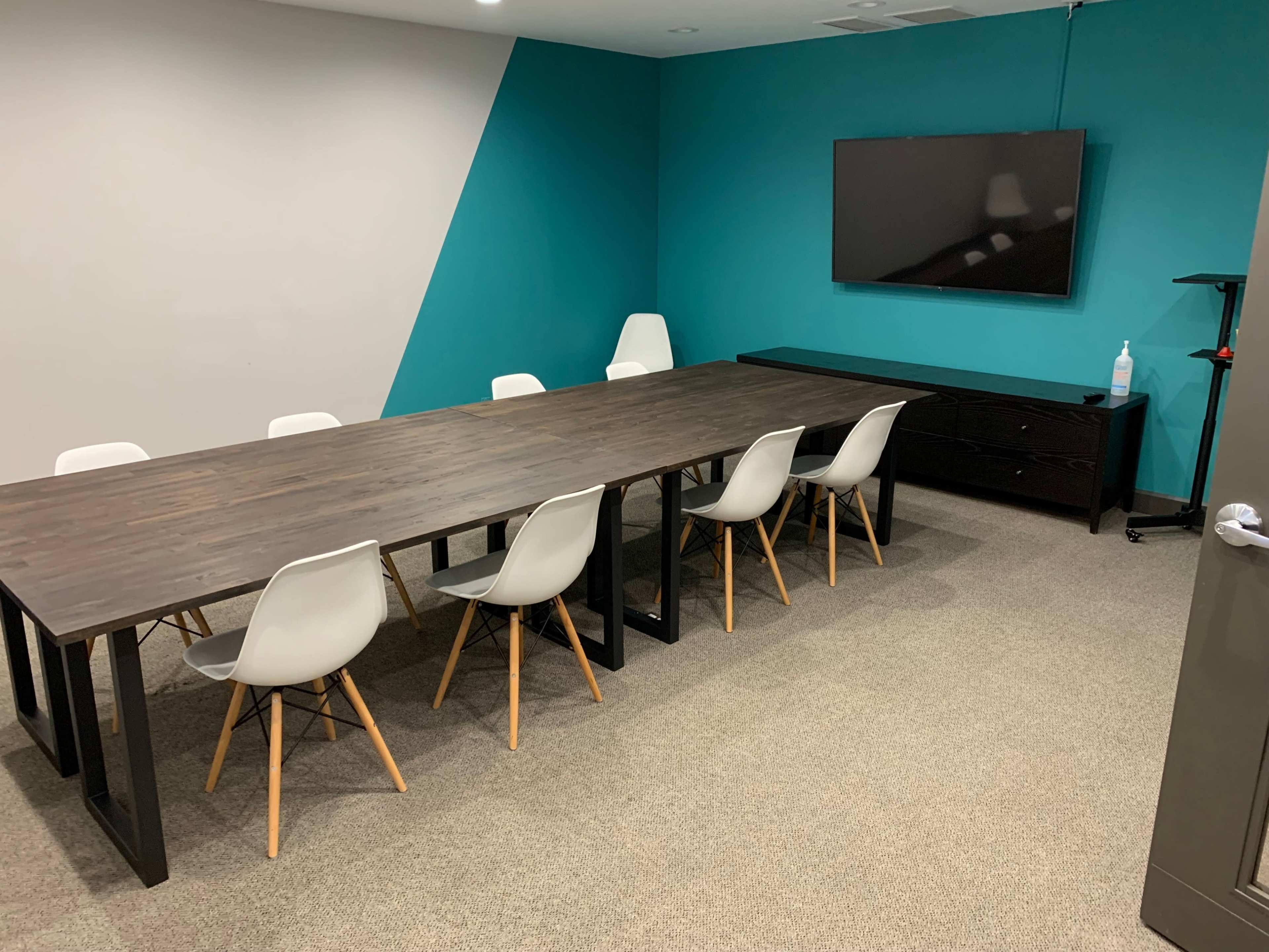 The image shows a conference room with a long wooden table surrounded by white chairs, a television mounted on the wall, and a side cabinet.