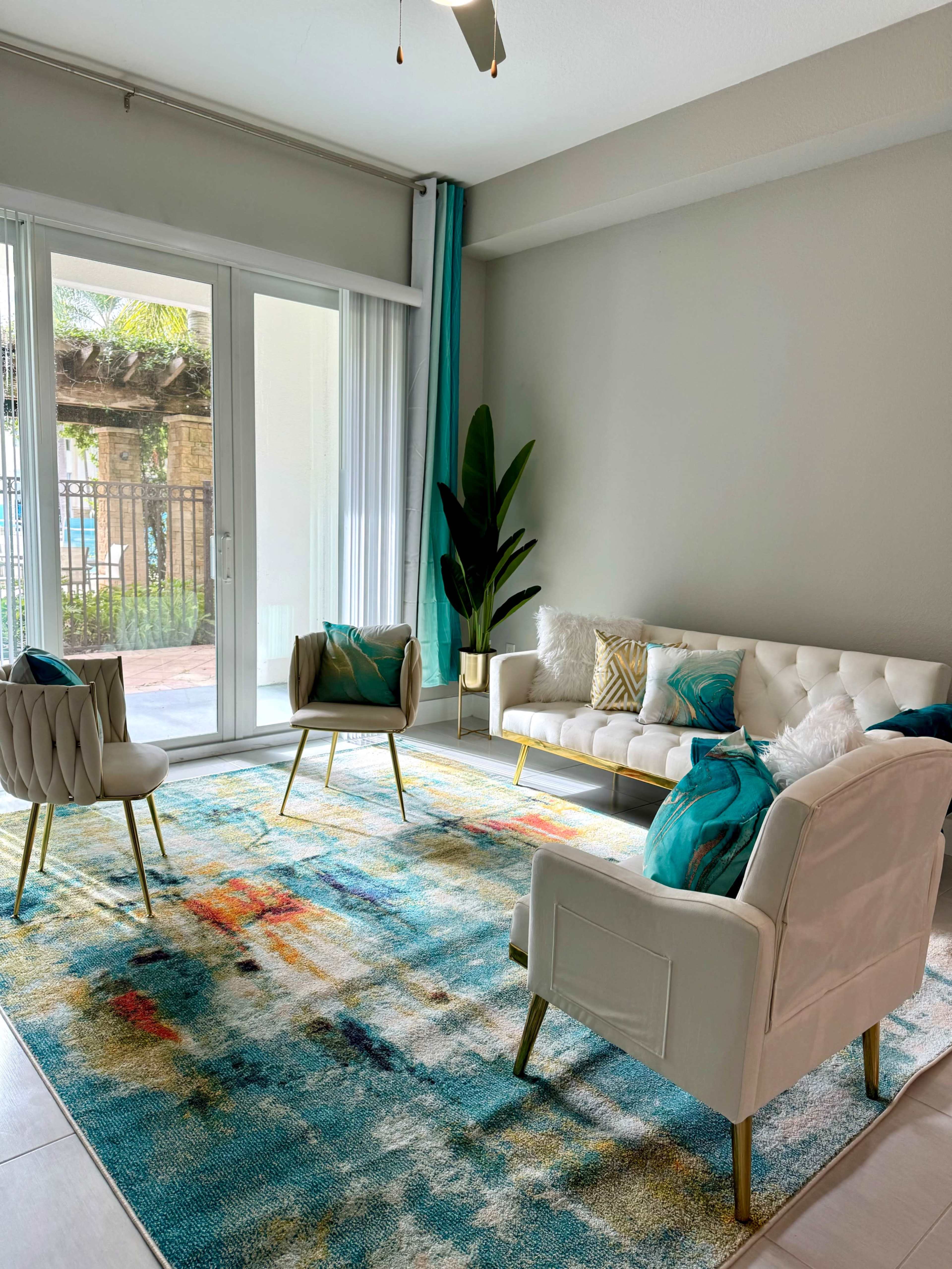 The image shows a modern living room with a white leather sofa, two tufted chairs, and a colorful rug, set against sliding glass doors that lead to a patio area.