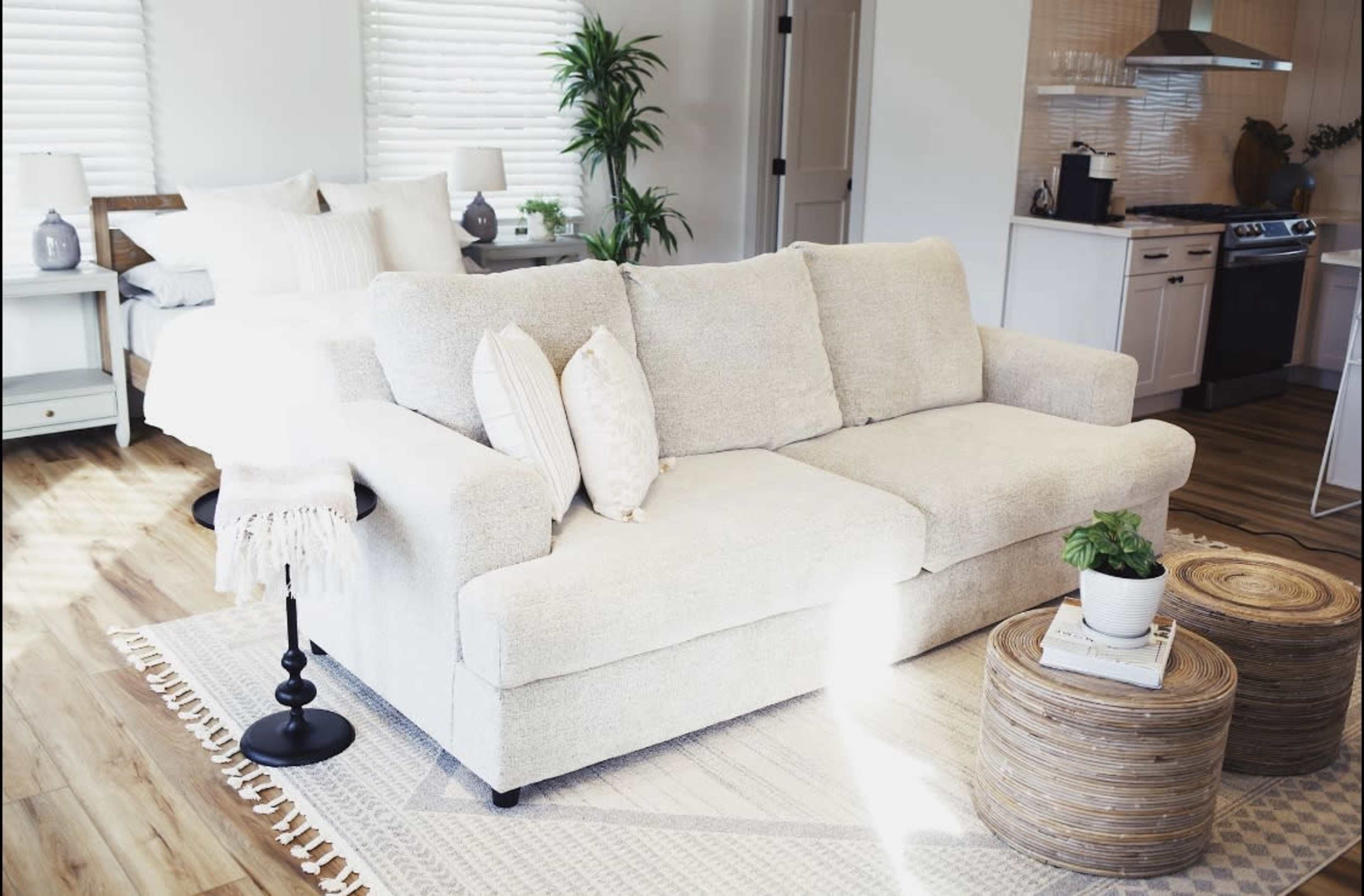 A light-colored sectional sofa is positioned in a cozy room with a bed in the background, accompanied by a wooden coffee table and a small plant.