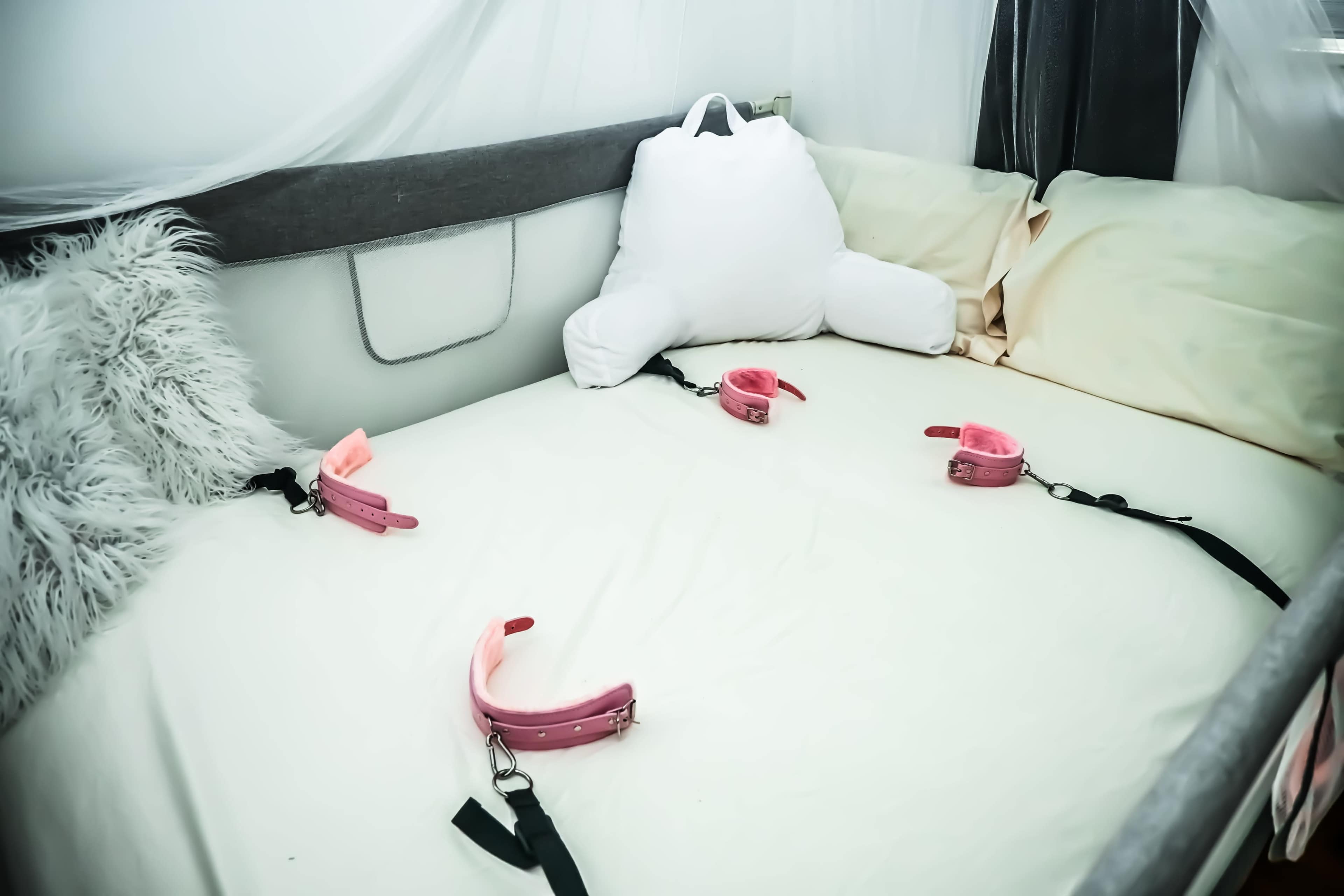 A bed with restraints positioned at each corner and a large pillow in the shape of a stuffed animal lies in the center.