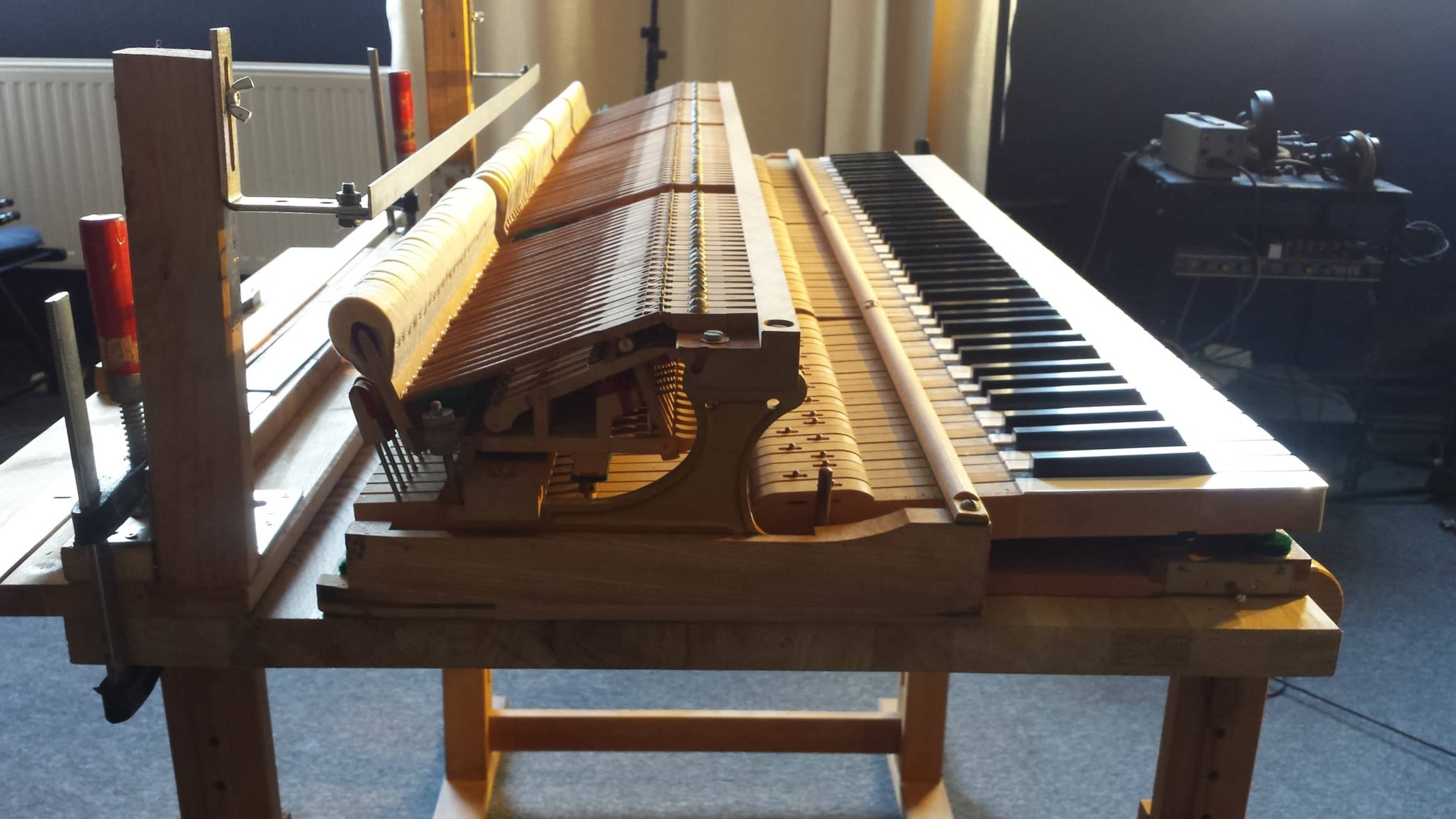 An open grand piano with visible internal mechanisms and keys is positioned on a wooden stand in a well-lit room.