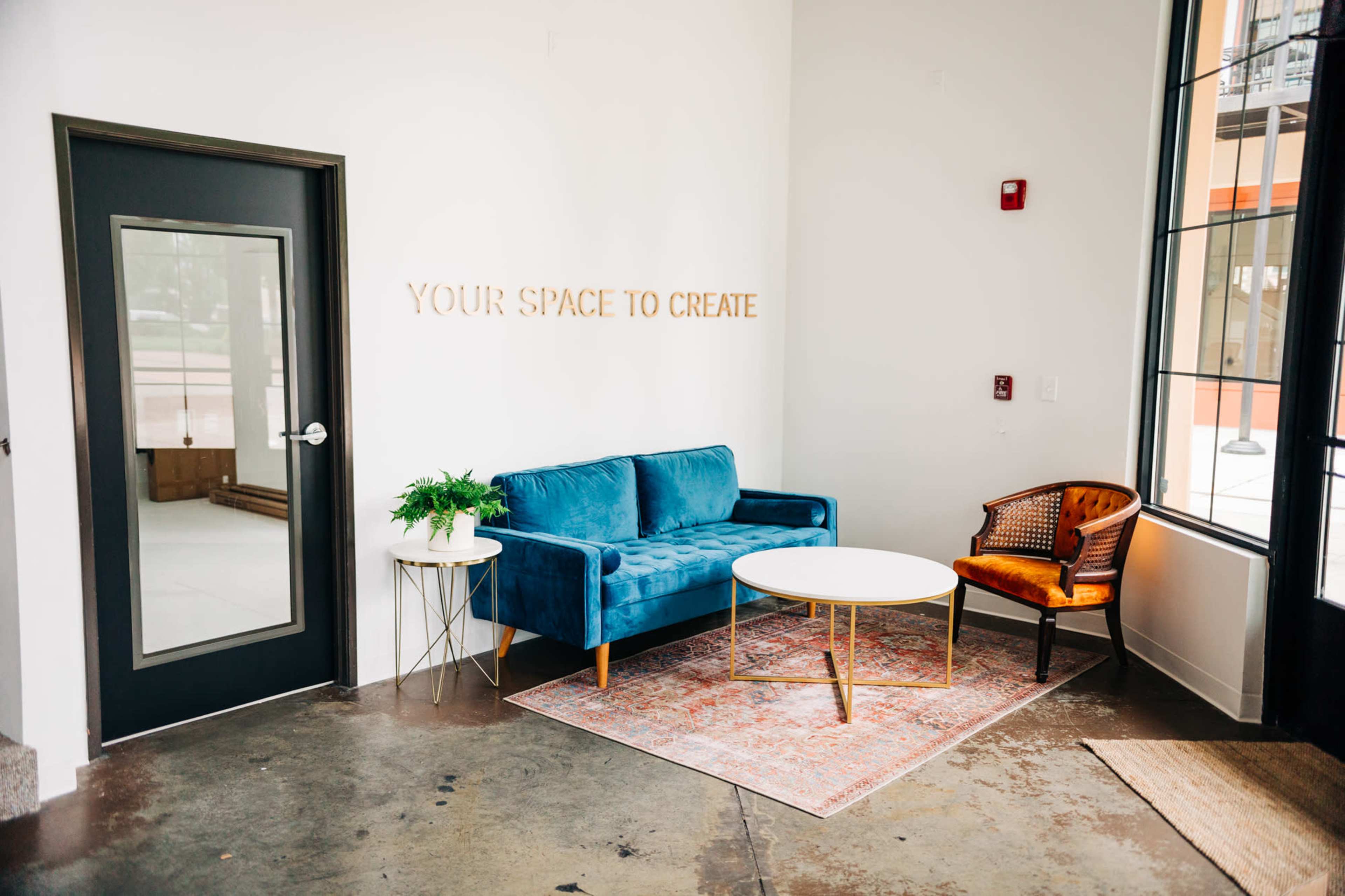 A blue velvet sofa with a small round table is positioned in a modern lobby area featuring minimalistic decor and large windows.