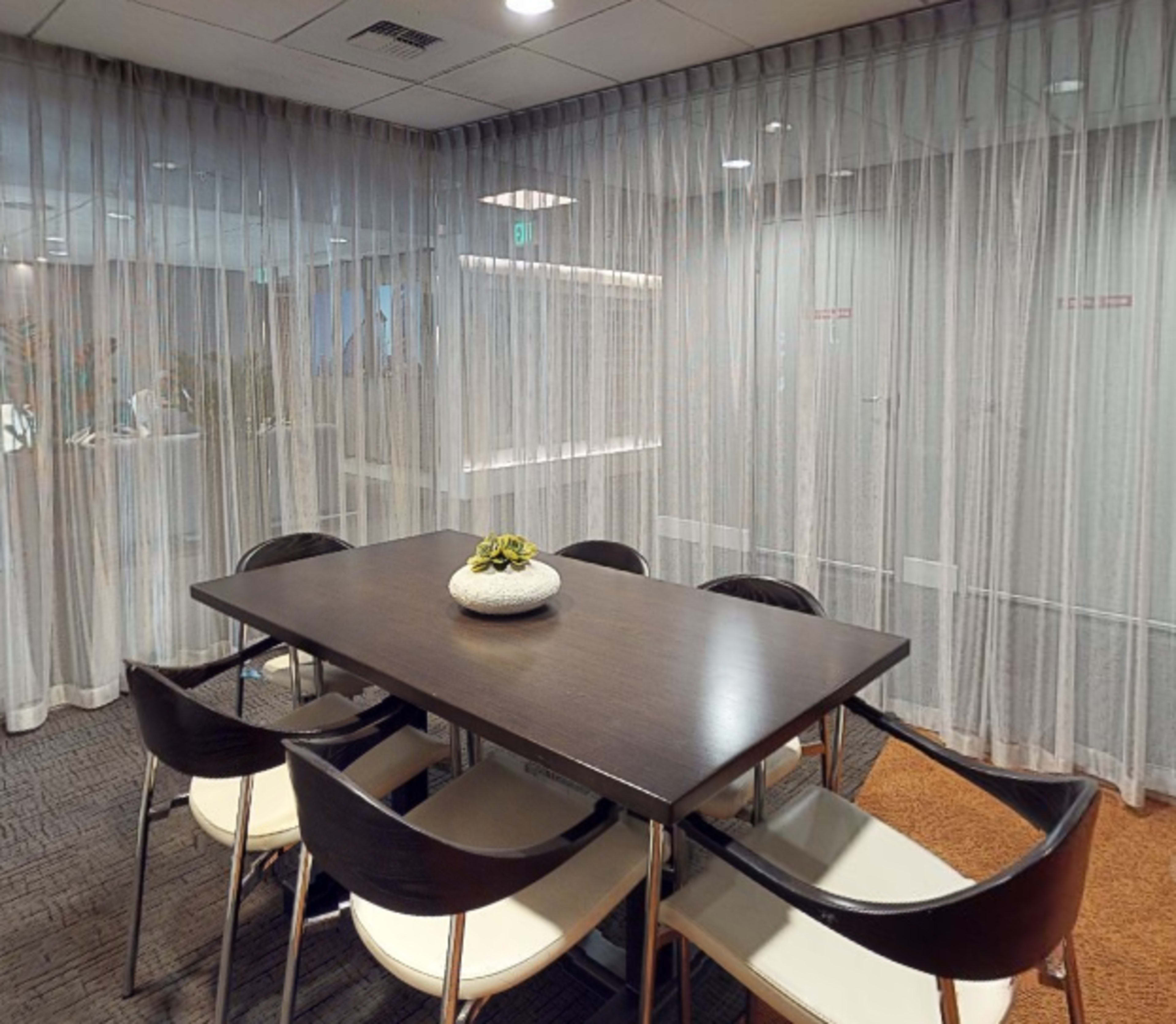 A conference room with a dark wooden table surrounded by white chairs, enclosed by sheer curtain panels.