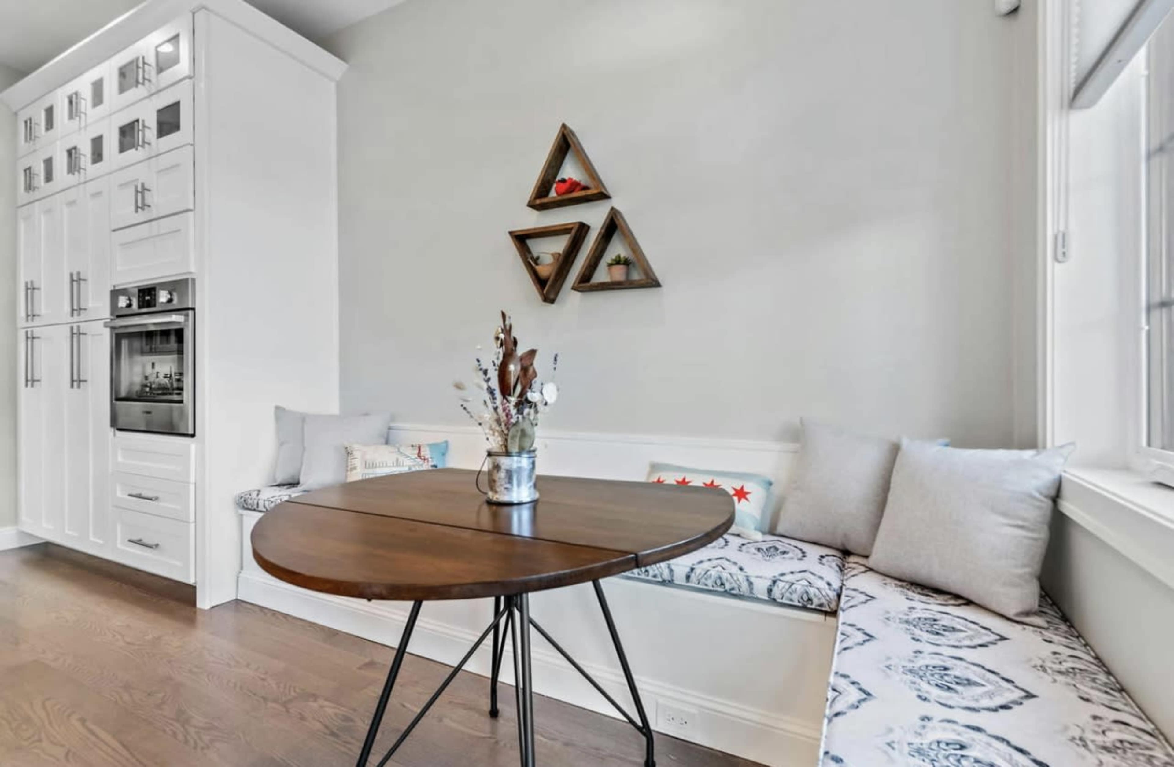 A round wooden table sits in front of a built-in bench with decorative pillows against a light-colored wall featuring triangular wall art.