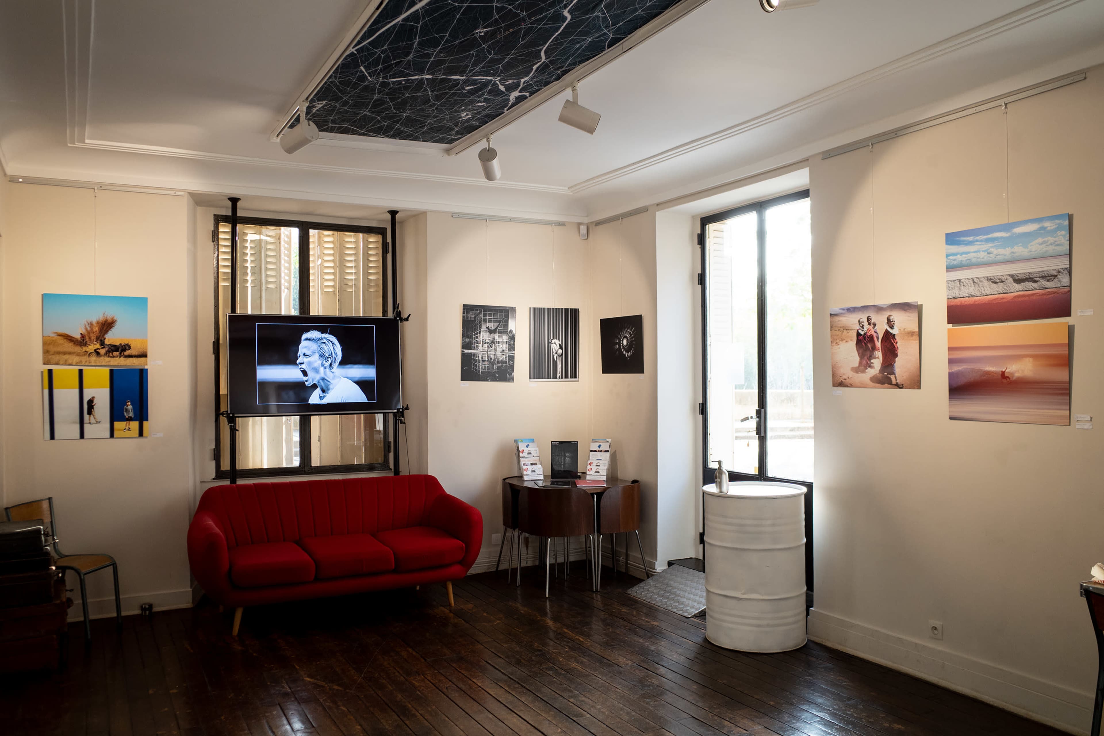 The image depicts an art gallery featuring various photographs displayed on the walls, a red couch in the foreground, and a TV screen showing an image.