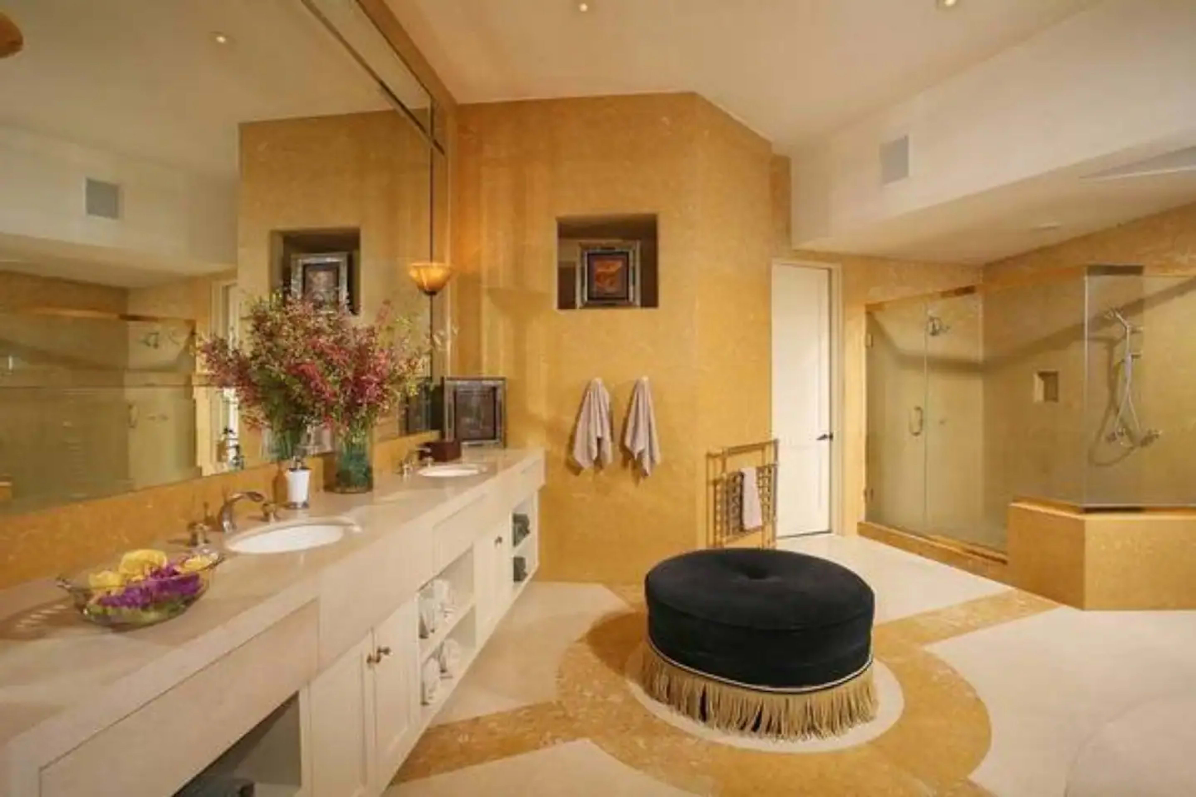 The bathroom features a large circular black ottoman, dual sinks with a mirror above, and a walk-in shower surrounded by golden-hued walls.