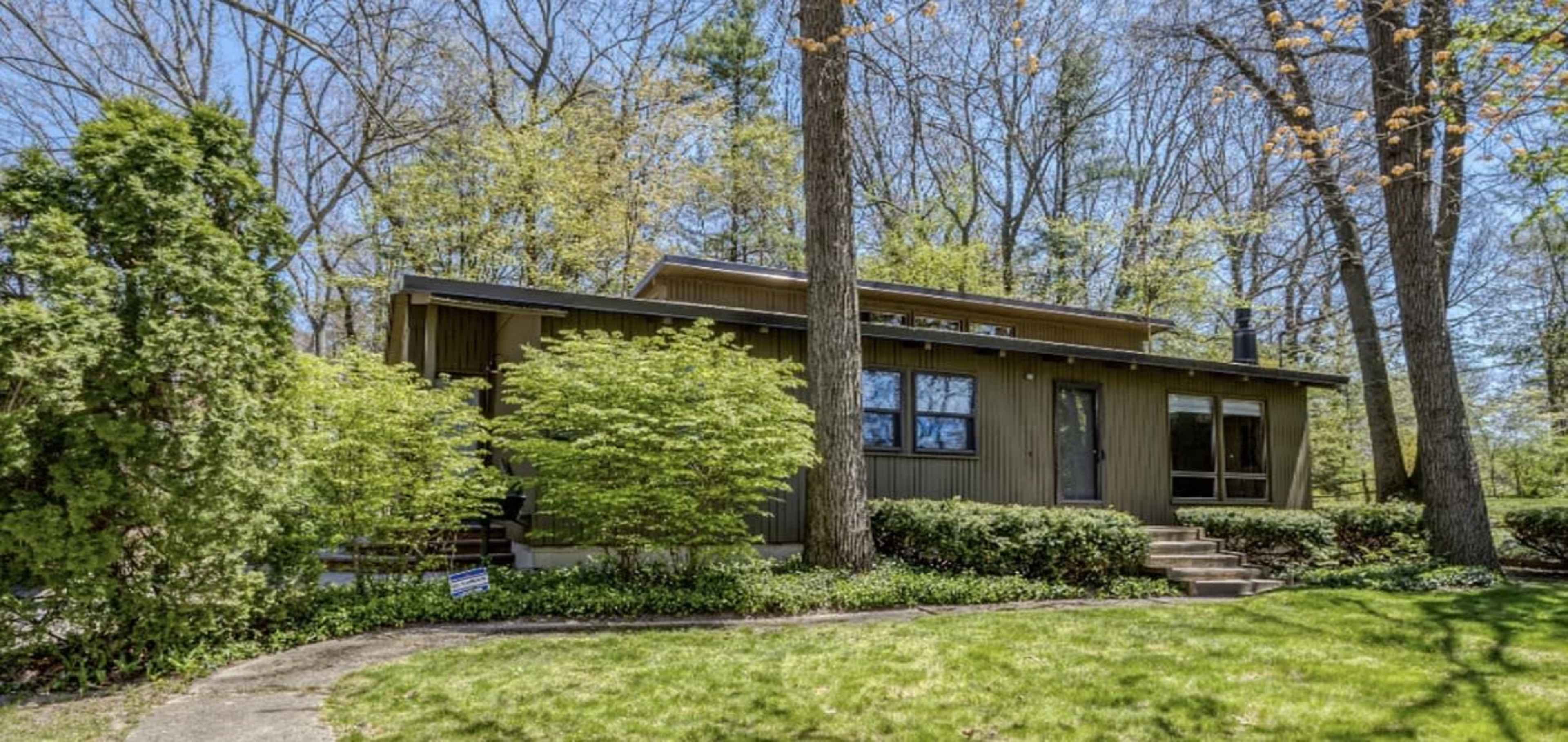 A single-story wooden house with large windows is surrounded by lush green grass and trees.